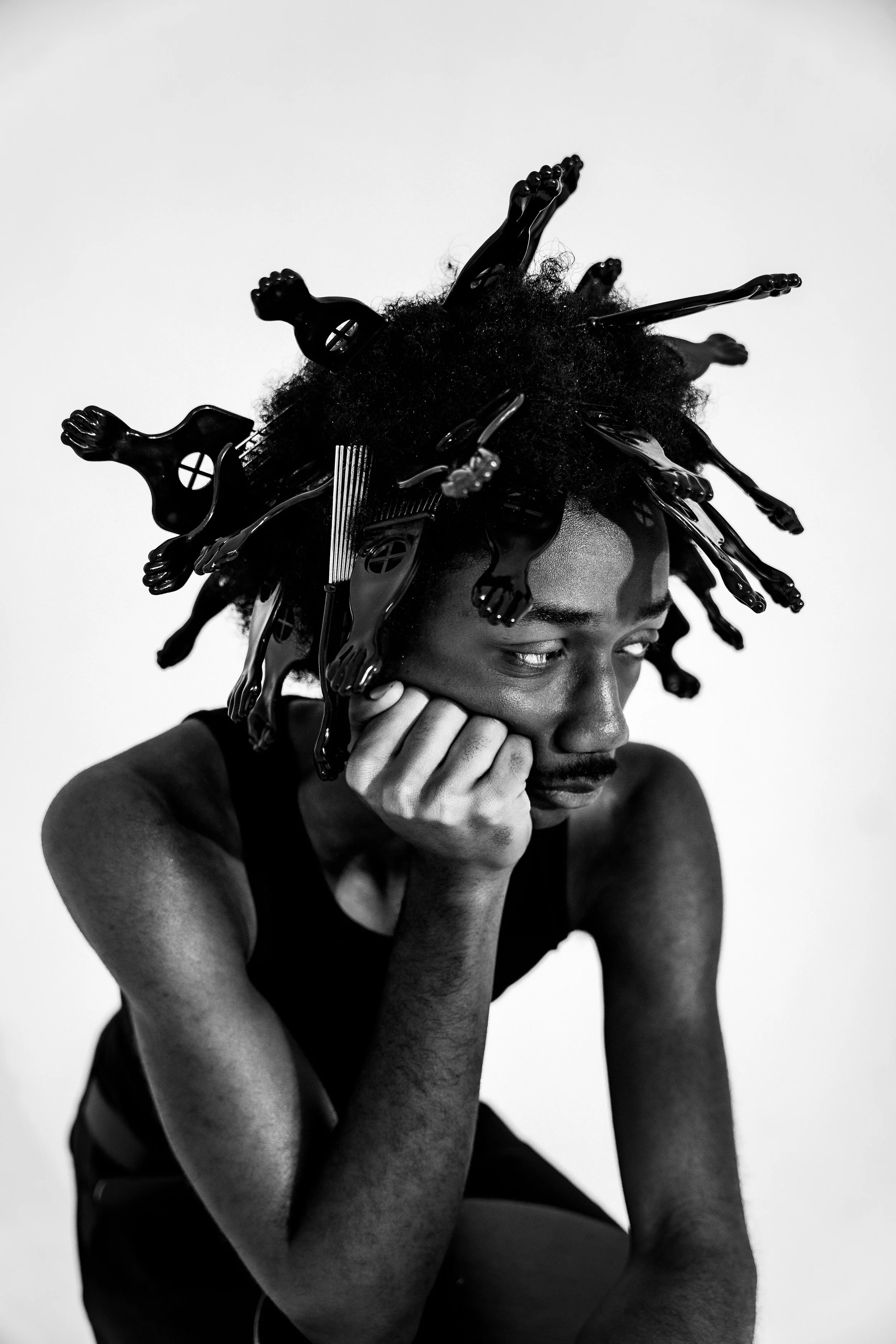 Black and white photo of a person with curly hair styled with numerous plastic hair clips, resting their chin on their hand and looking to the side.
