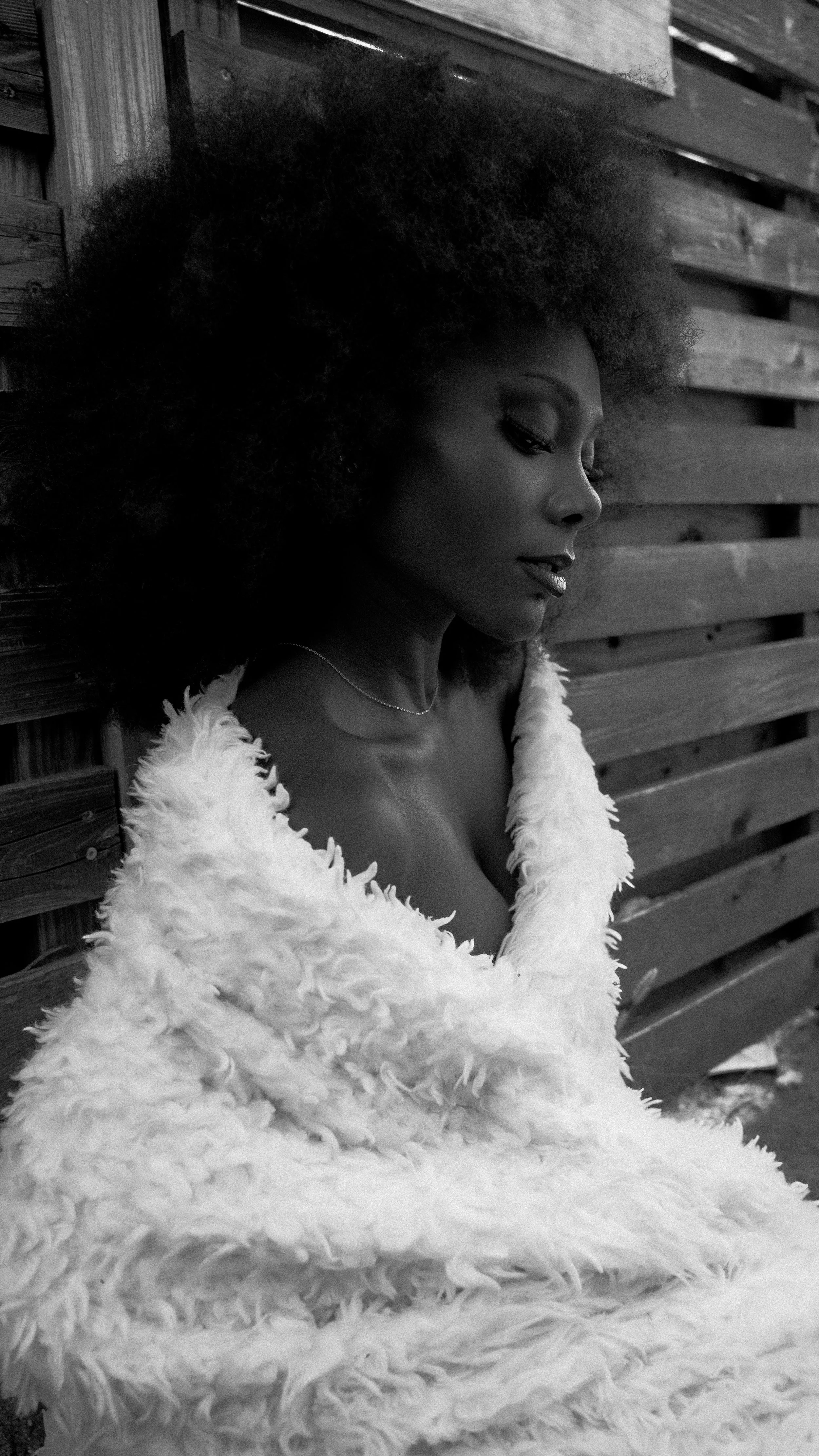 A black-and-white photo of a woman with voluminous curly hair, wearing a fluffy fur coat, sitting against a wooden fence, looking down thoughtfully.