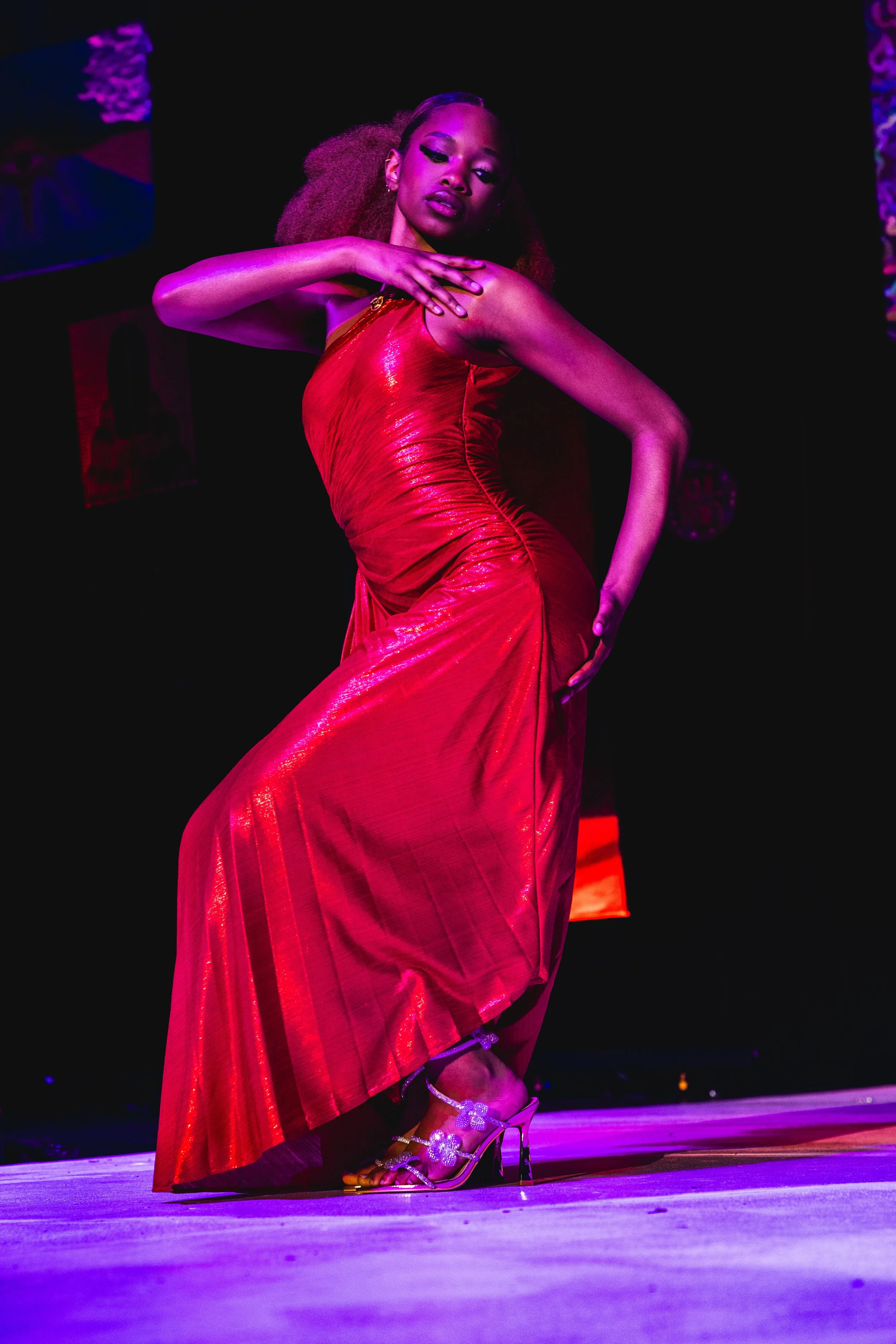A woman in a shiny red gown with a slit, performing a dance on stage with purple and pink lighting, wearing high heels decorated with flowers.