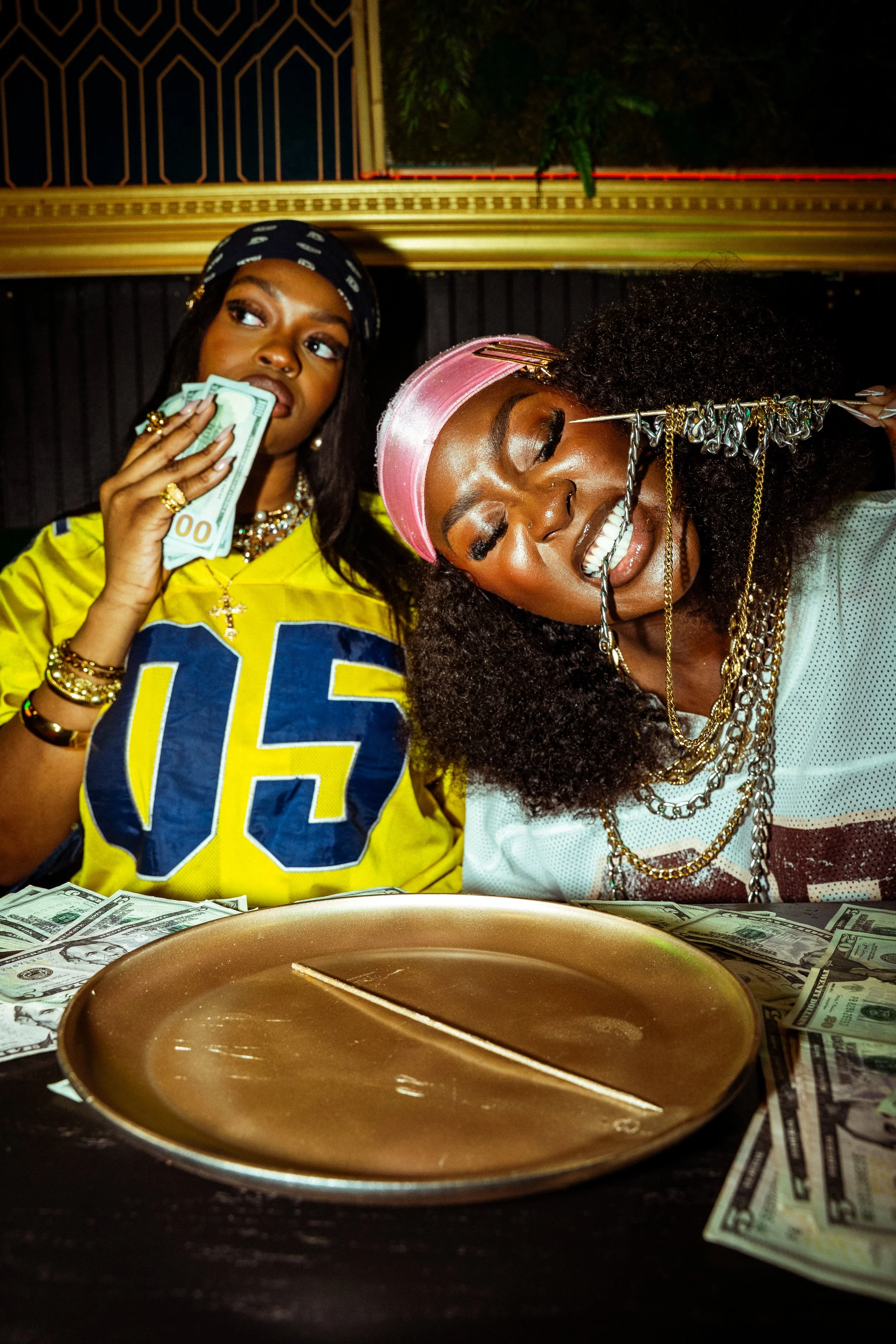 Two women with jewelry, one holding money and the other biting a chain, sitting at a table with money and a gold plate.
