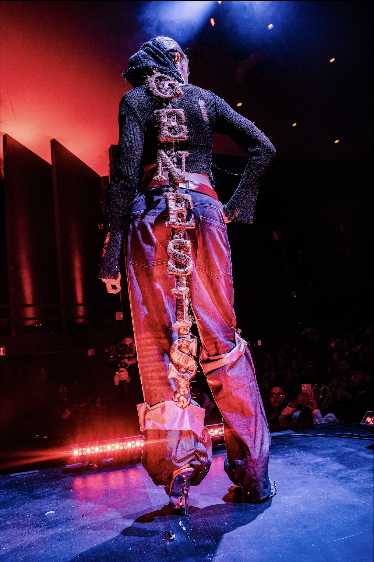 Back view of a runway model wearing a black sweater with large letters spelling 'LOVE' down the back, high-waisted baggy jeans with a chain, clear high heels, on a stage with red and blue lighting, and audience members visible in the background.