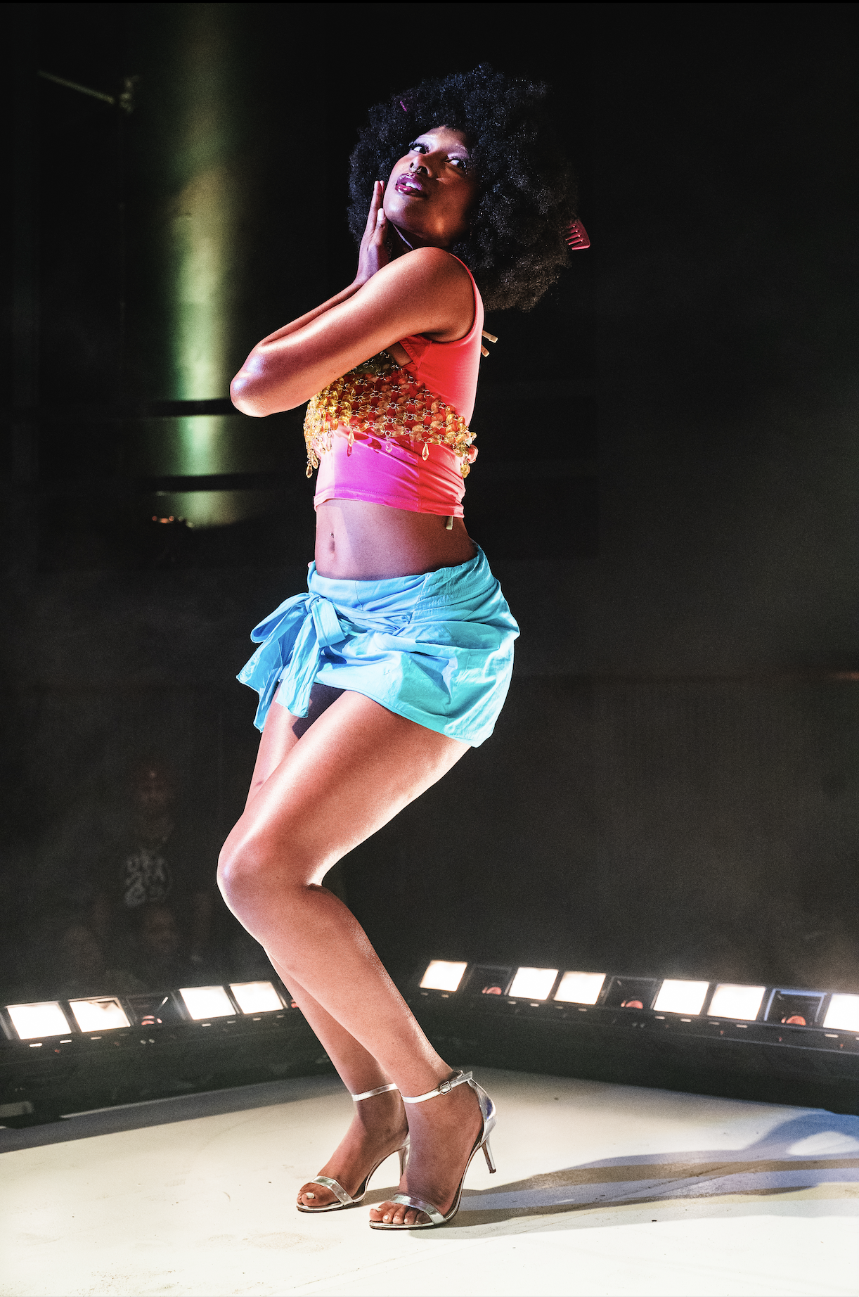 A woman with curly black hair dancing on stage, wearing a pink and gold top, blue shorts, and high heels.