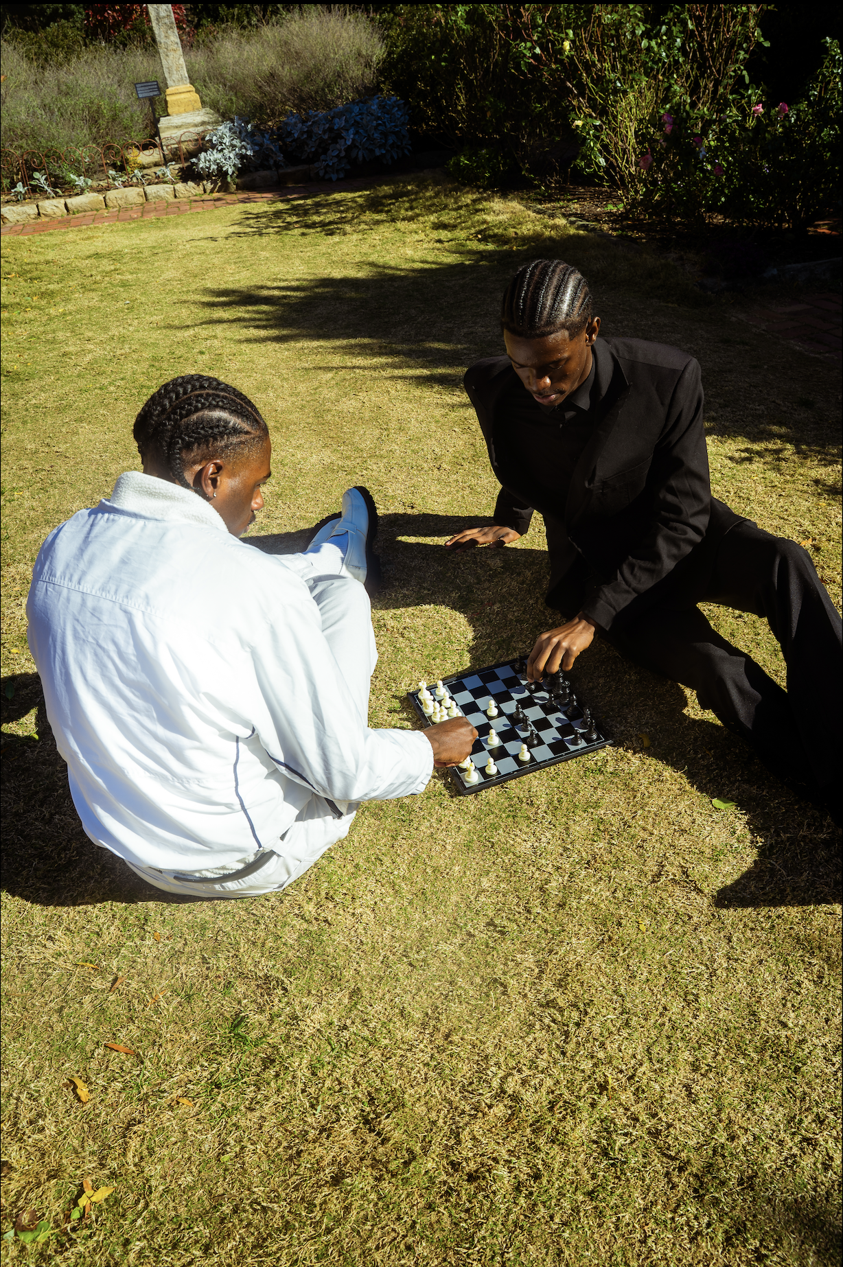 Two young men playing chess outdoors on the grass in a garden with bushes and flowers.