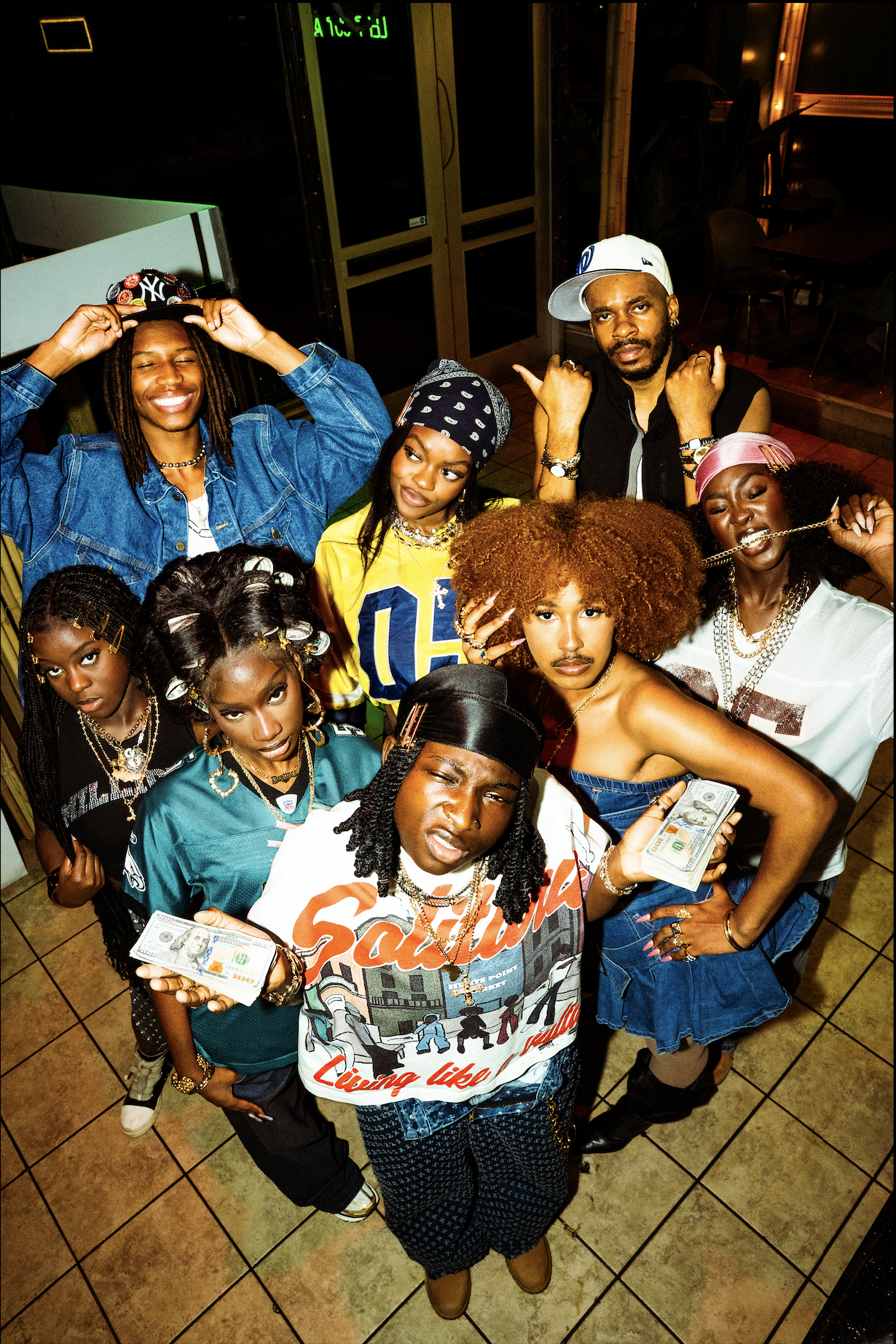 A group of ten people, mostly women, posing together in a nightlife setting. Some hold stacks of cash, and one person in the front left is wearing a sports jersey. They are dressed in casual, fashionable clothing with accessories, and the mood appear