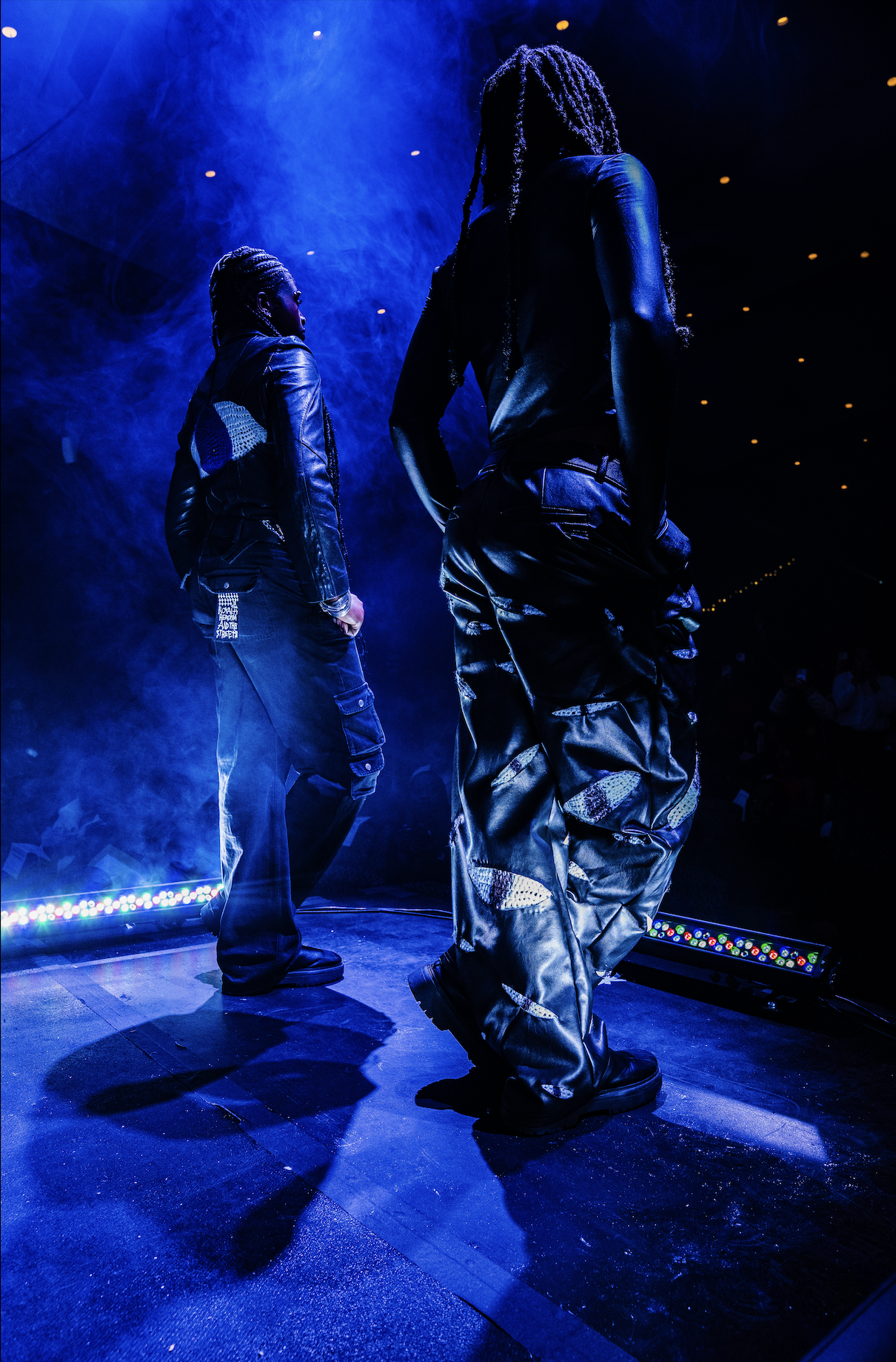 Two performers on stage under blue and purple lighting, dressed in black outfits with camouflage details, facing away from the camera. Smoke effects and stage lights create a dramatic atmosphere.