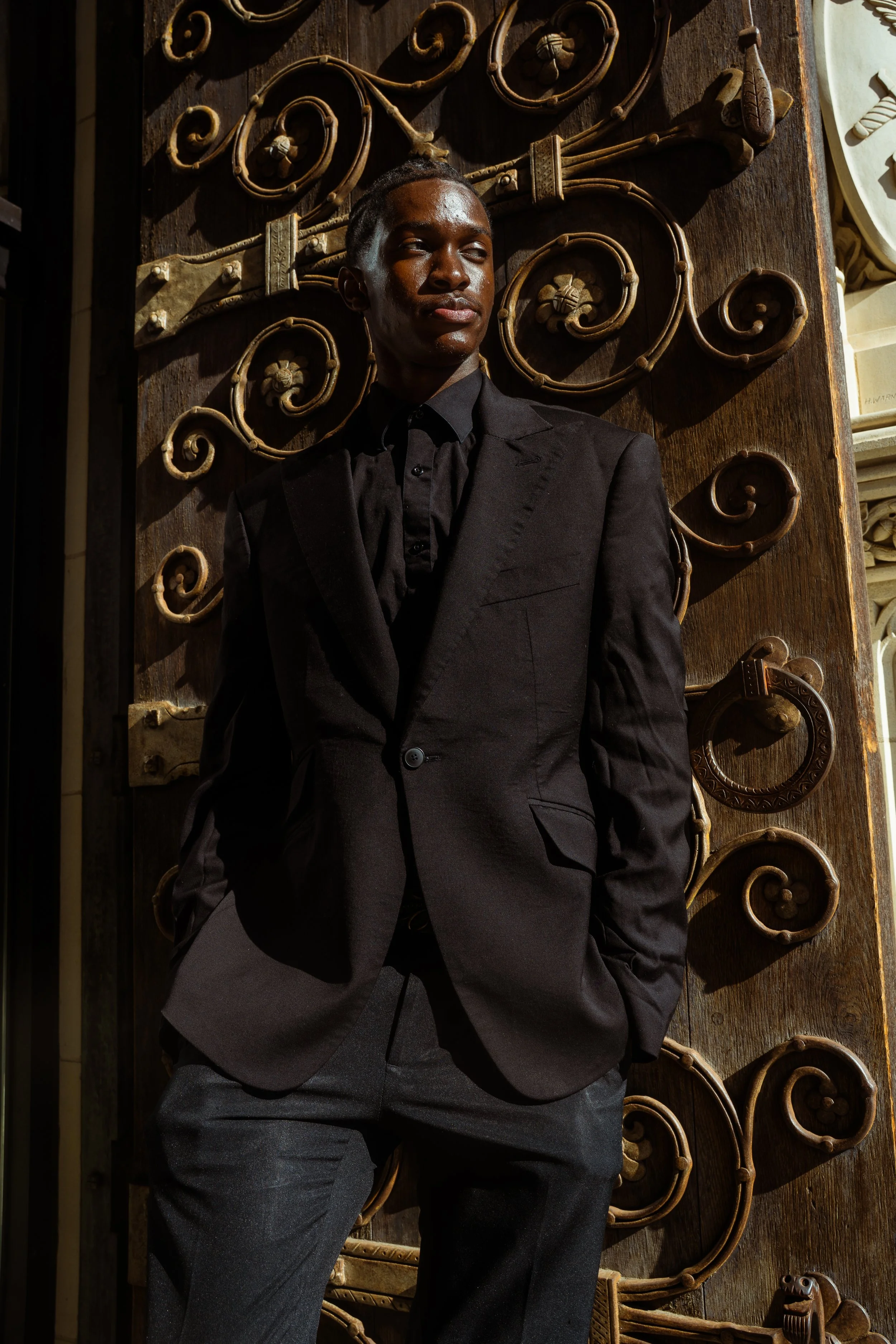 A person in a black suit and shirt stands against an ornate brown metal gate with gold accents, partially illuminated in warm lighting.