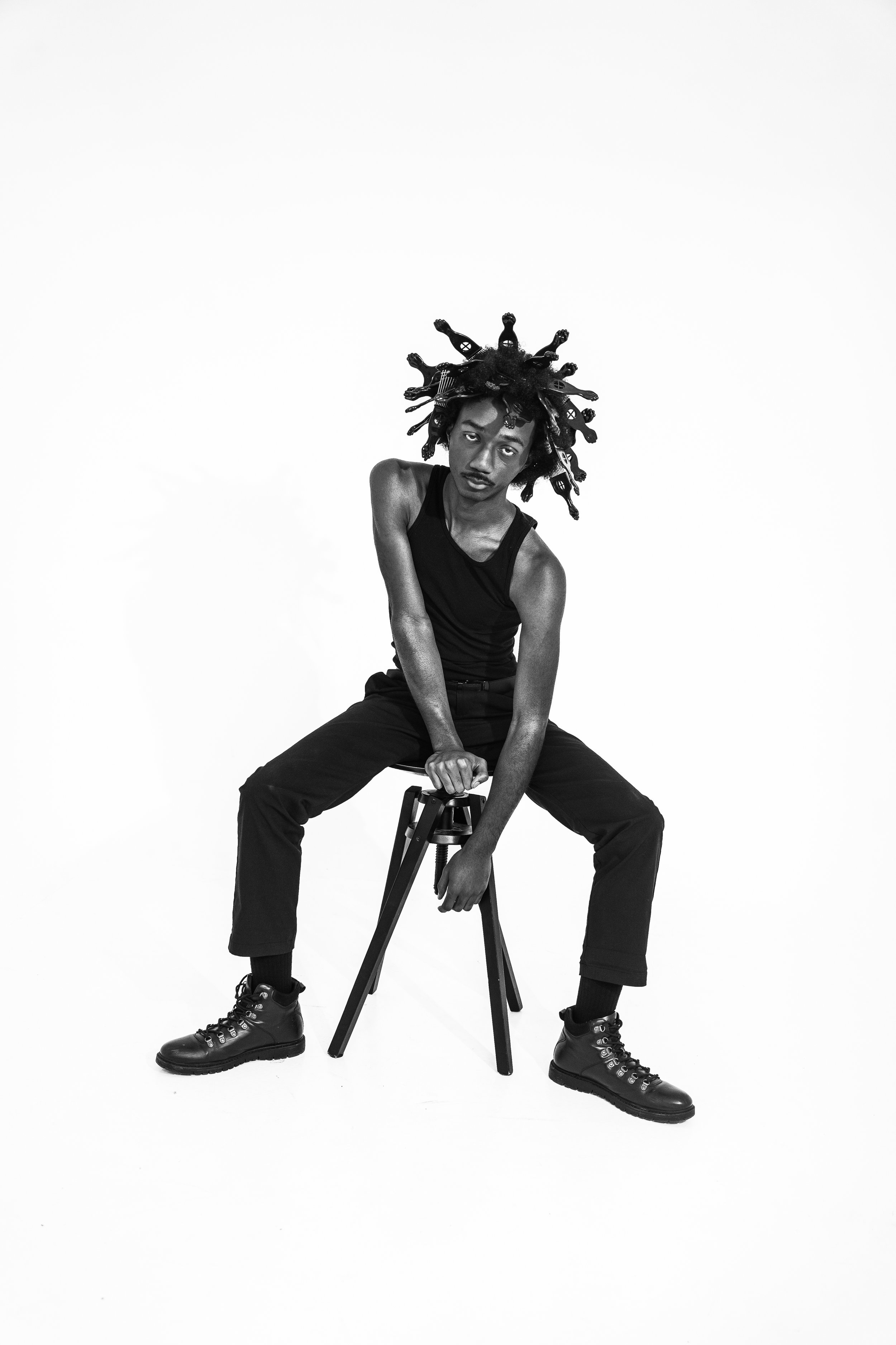 Black and white photo of a young man sitting on a stool with a plain white background. He has dreadlocks and is wearing a black tank top, black pants, and black boots. He has a serious expression and is leaning forward with one hand on his knee.