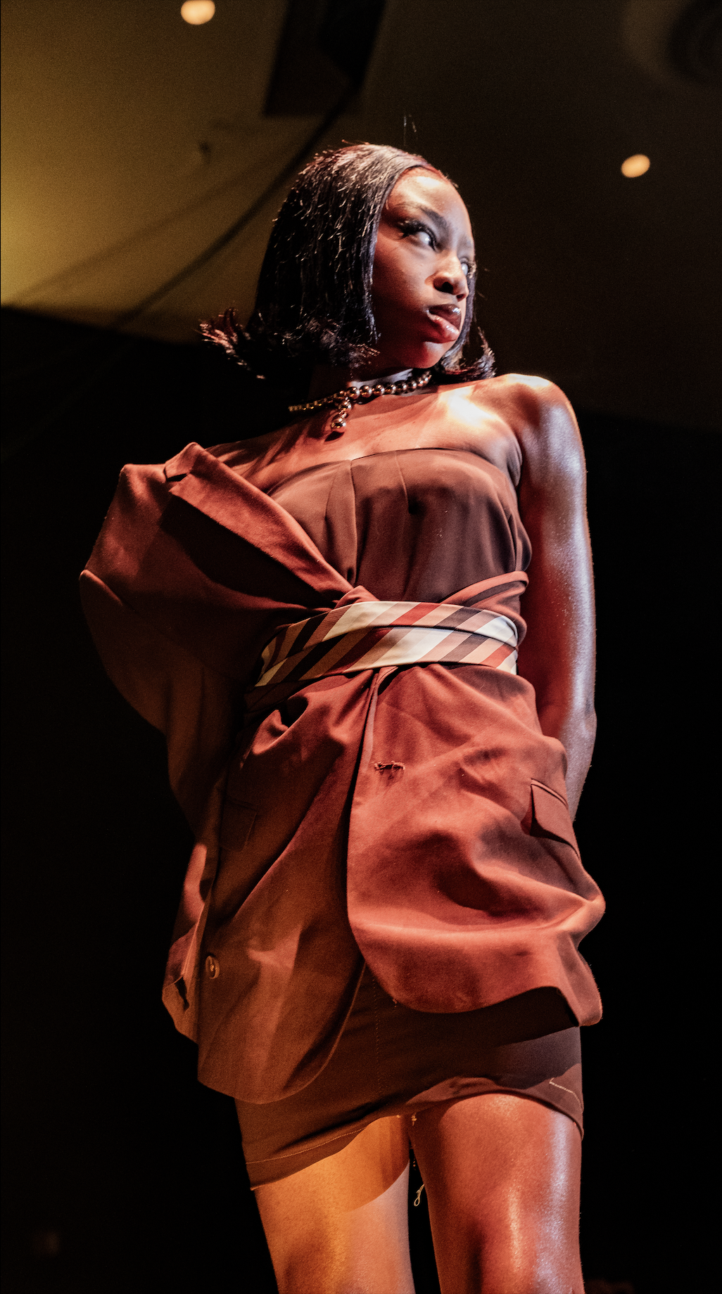 Fashion model walking on stage in a brown satin dress with a plaid belt, black hair styled in a bob, and a pearl necklace.