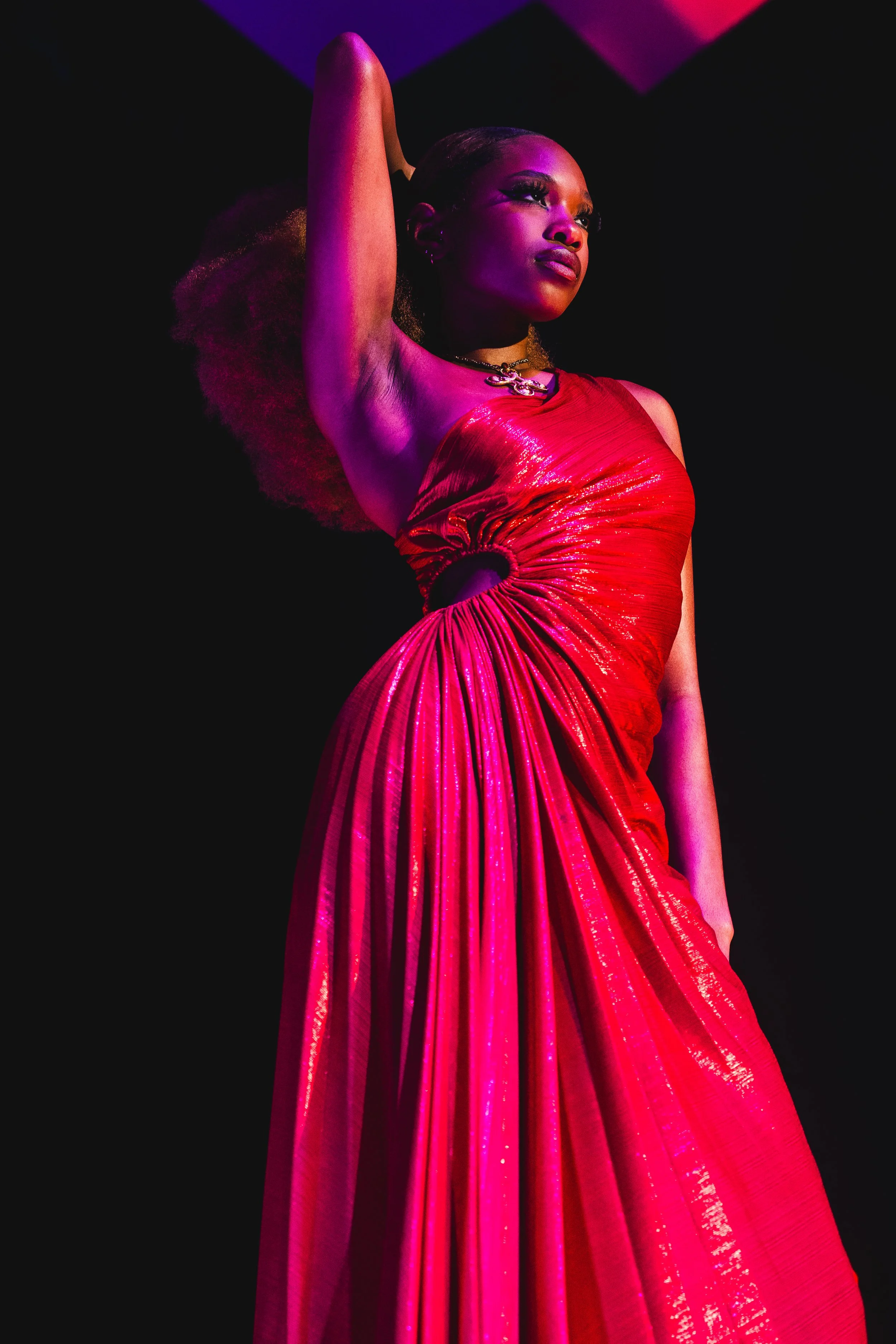 A woman wearing a shiny red dress with a large pleated skirt posing with one arm raised and resting behind her head against a dark background with pink and purple lights.