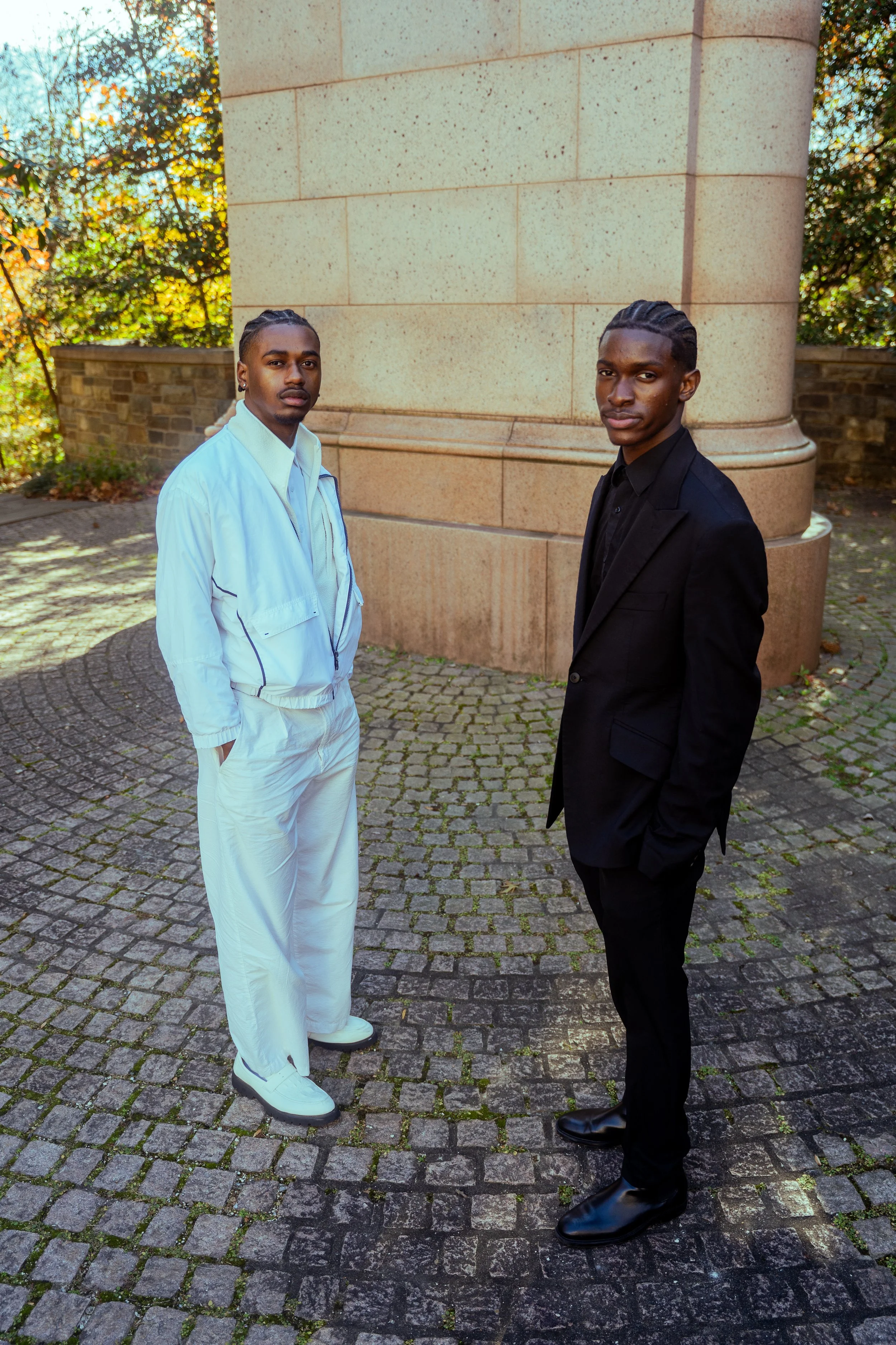 Two young men standing outdoors on a cobblestone path near a stone structure, one dressed in a white tracksuit and the other in a black suit, with autumn trees in the background.