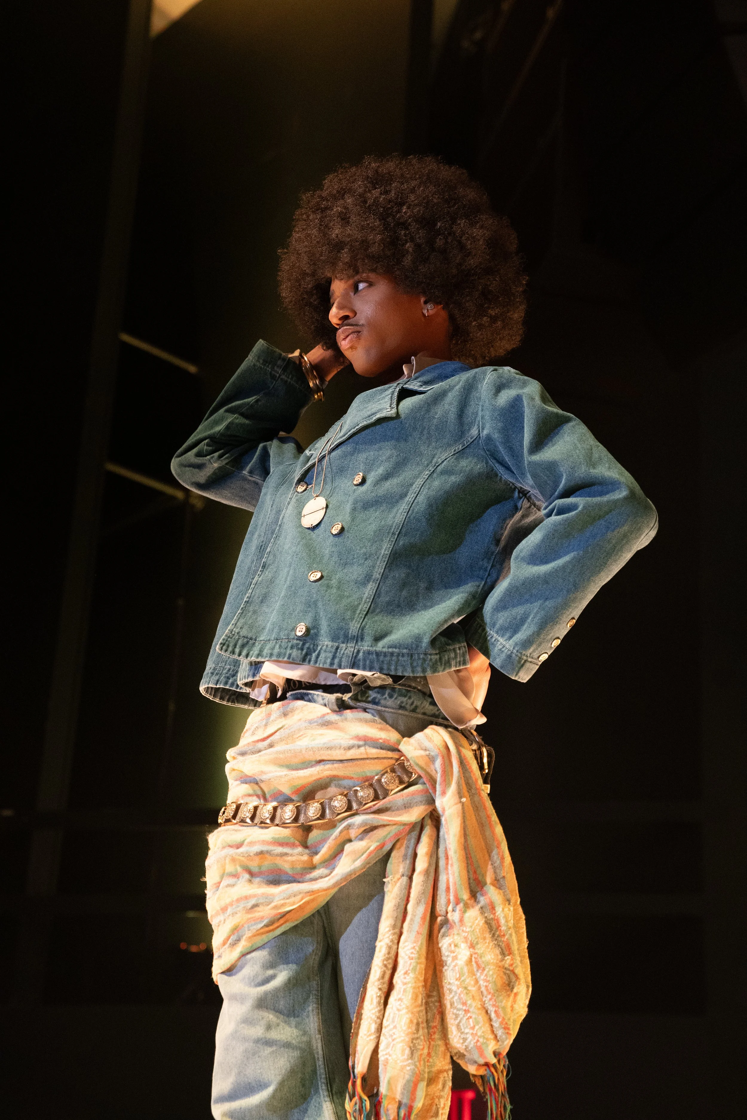 A woman with an afro hairstyle stands with her hand behind her head, wearing a denim jacket, a necklace, a colorful scarf tied around her waist, and jeans, in a dimly lit room.