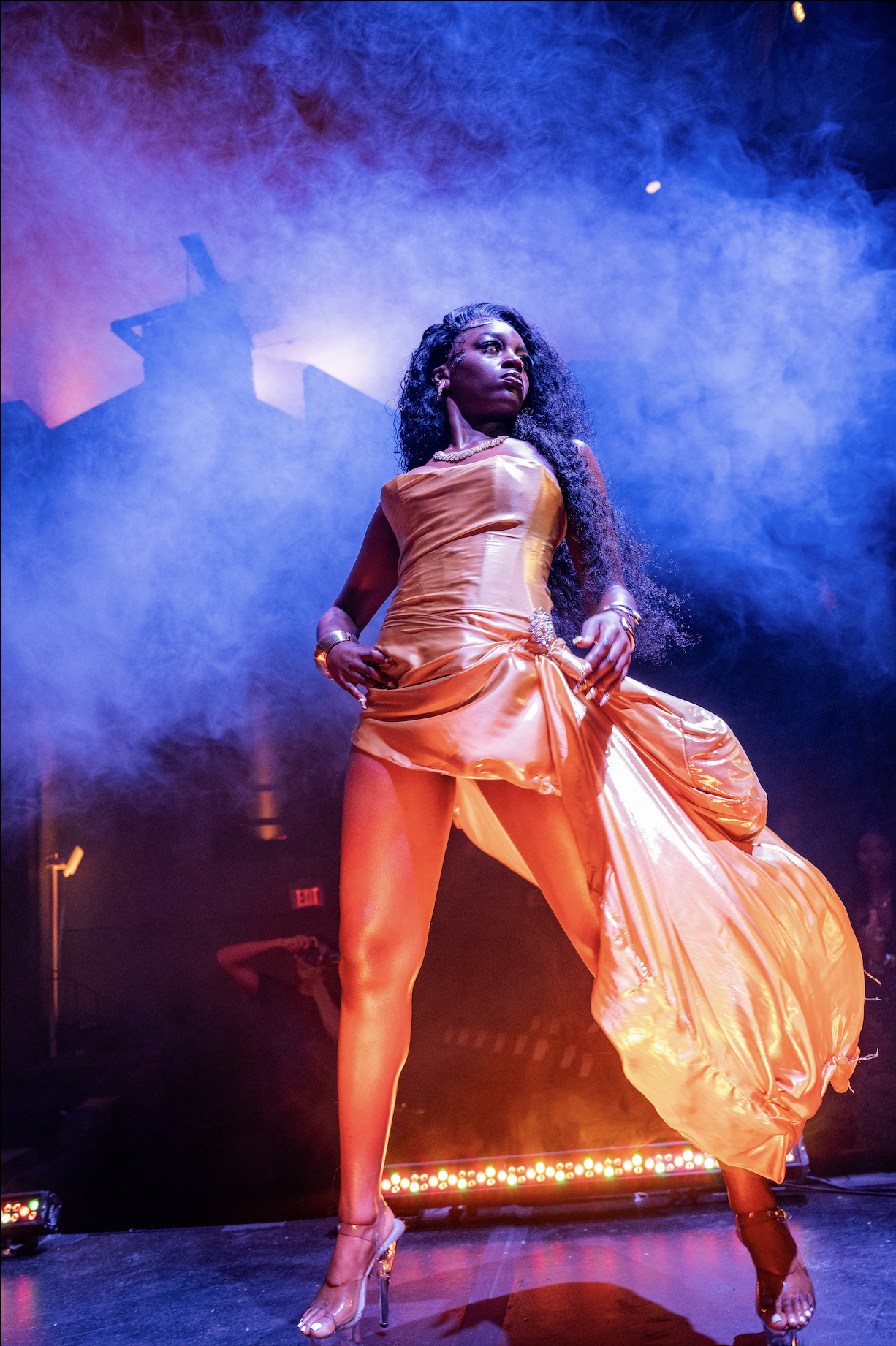 A woman in an elegant orange satin dress poses on stage with colorful lighting and smoke effects.