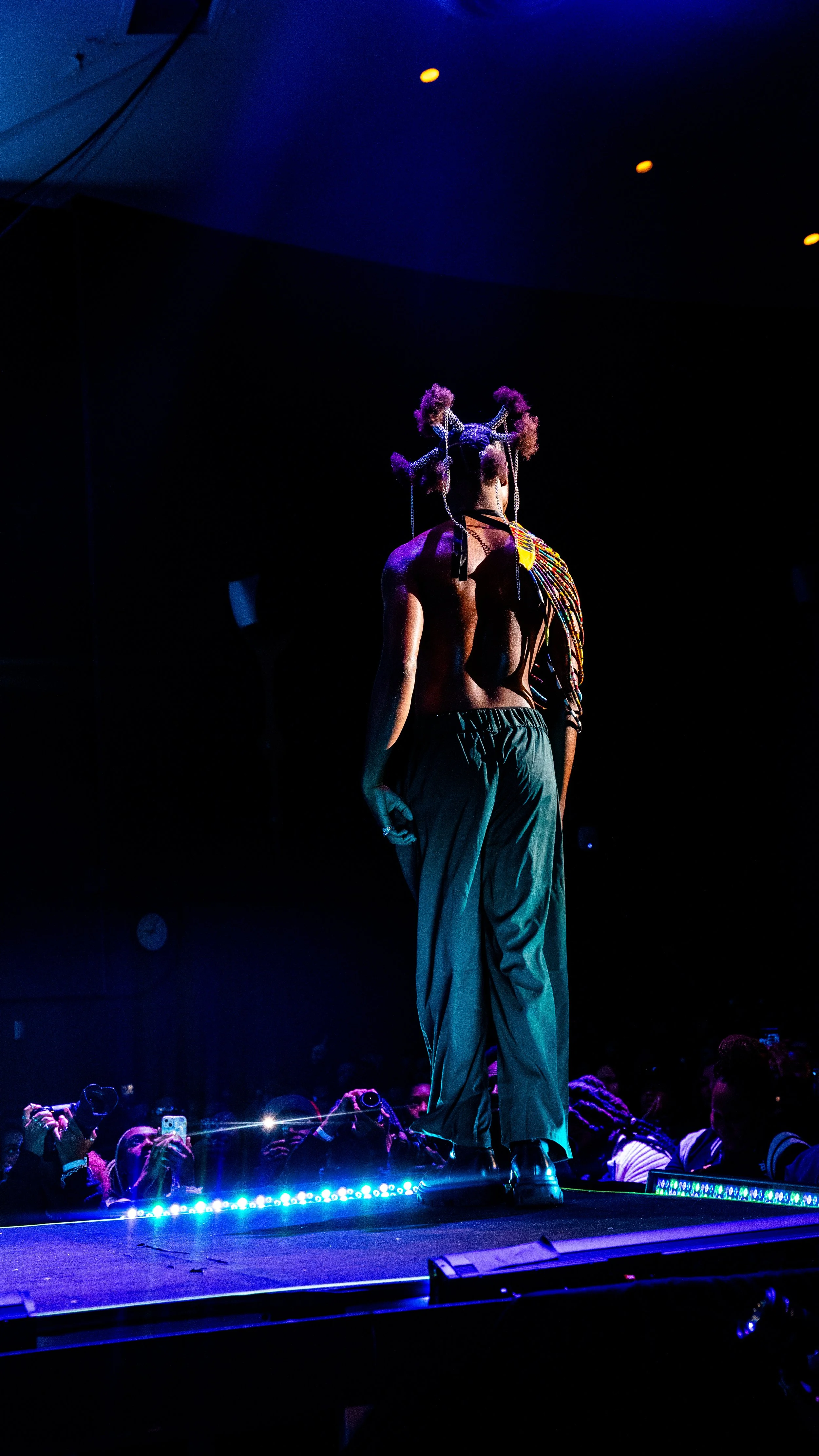 A model walking on a fashion runway with a dark background, wearing loose green pants, a black top, and an elaborate headpiece with colorful accessories.