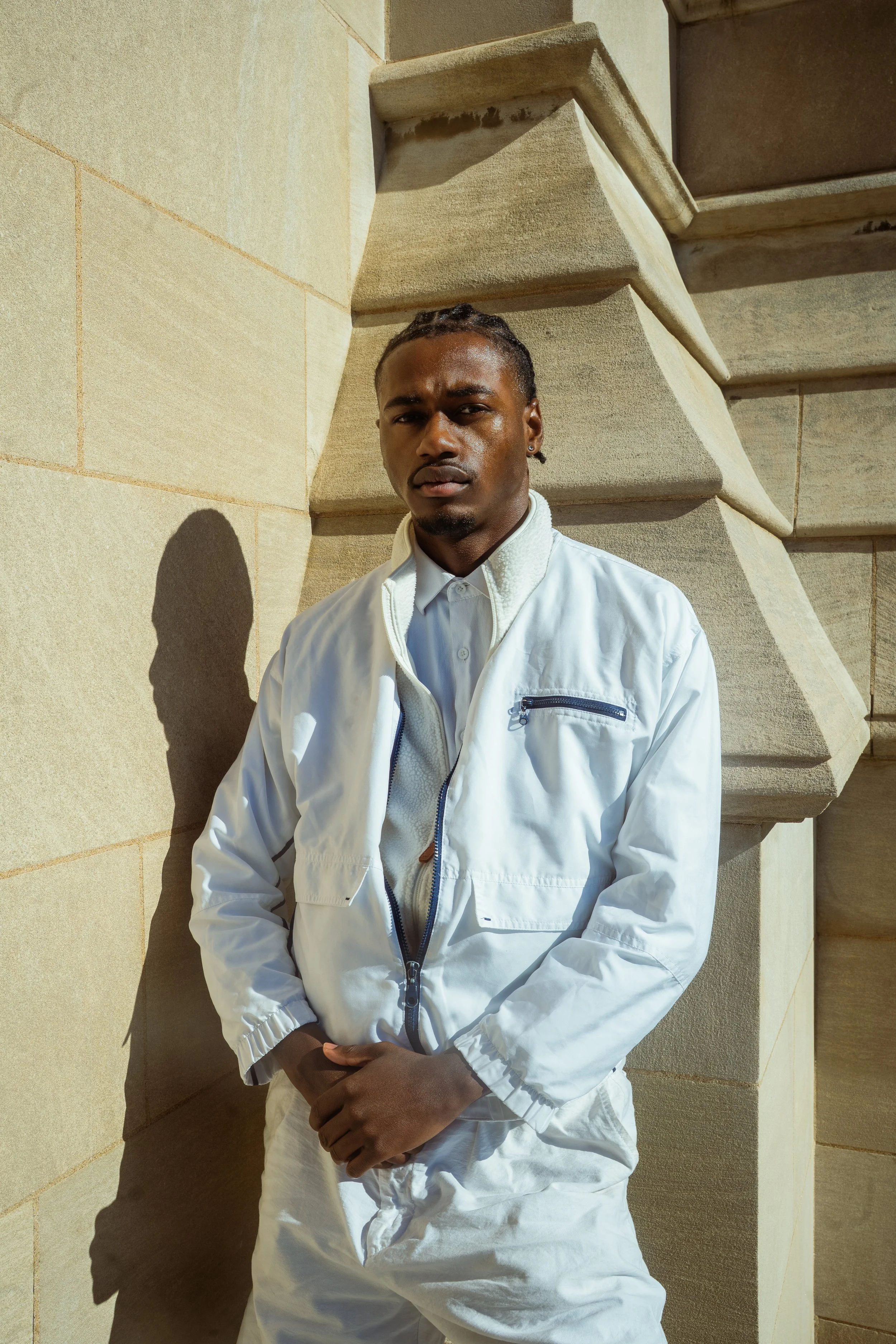 A young man with dark skin and braided hair stands outdoors against a stone wall and architectural ledge, casting a shadow. He is wearing a white jacket over a white shirt.
