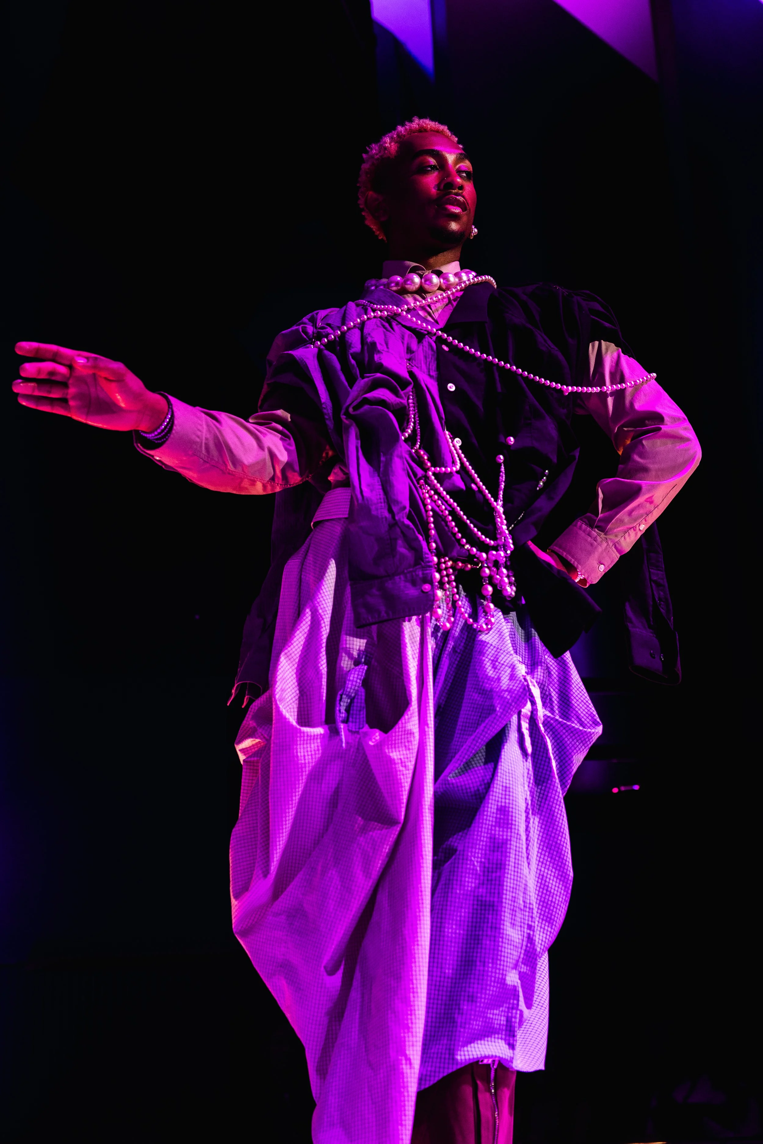 Person with short blonde curly hair wearing layered clothing and pearl necklaces, standing with one hand extended against a dark background with purple lighting.