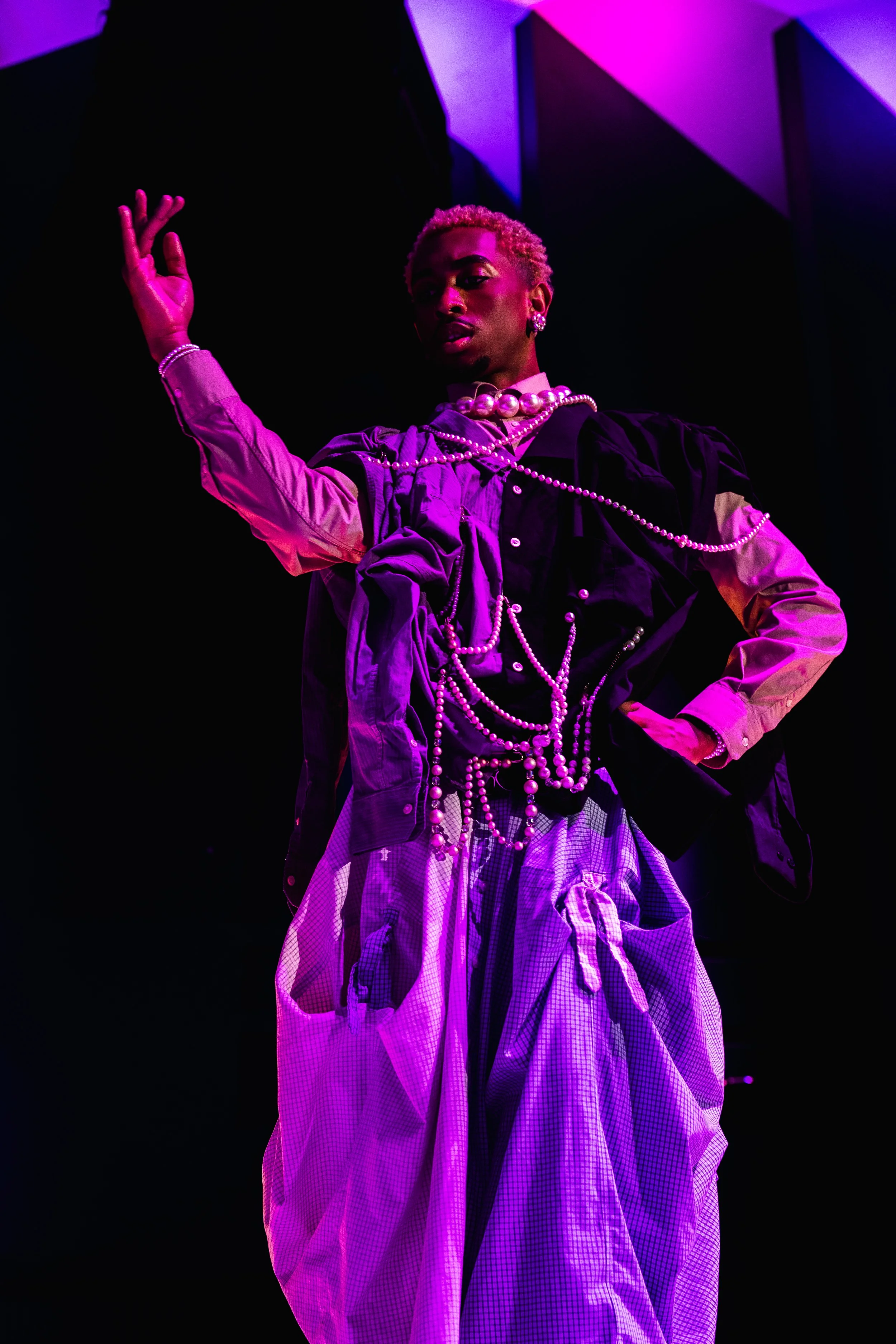 Person wearing layered clothing, jewelry, and patterned pants, with a hand raised, under purple lighting.