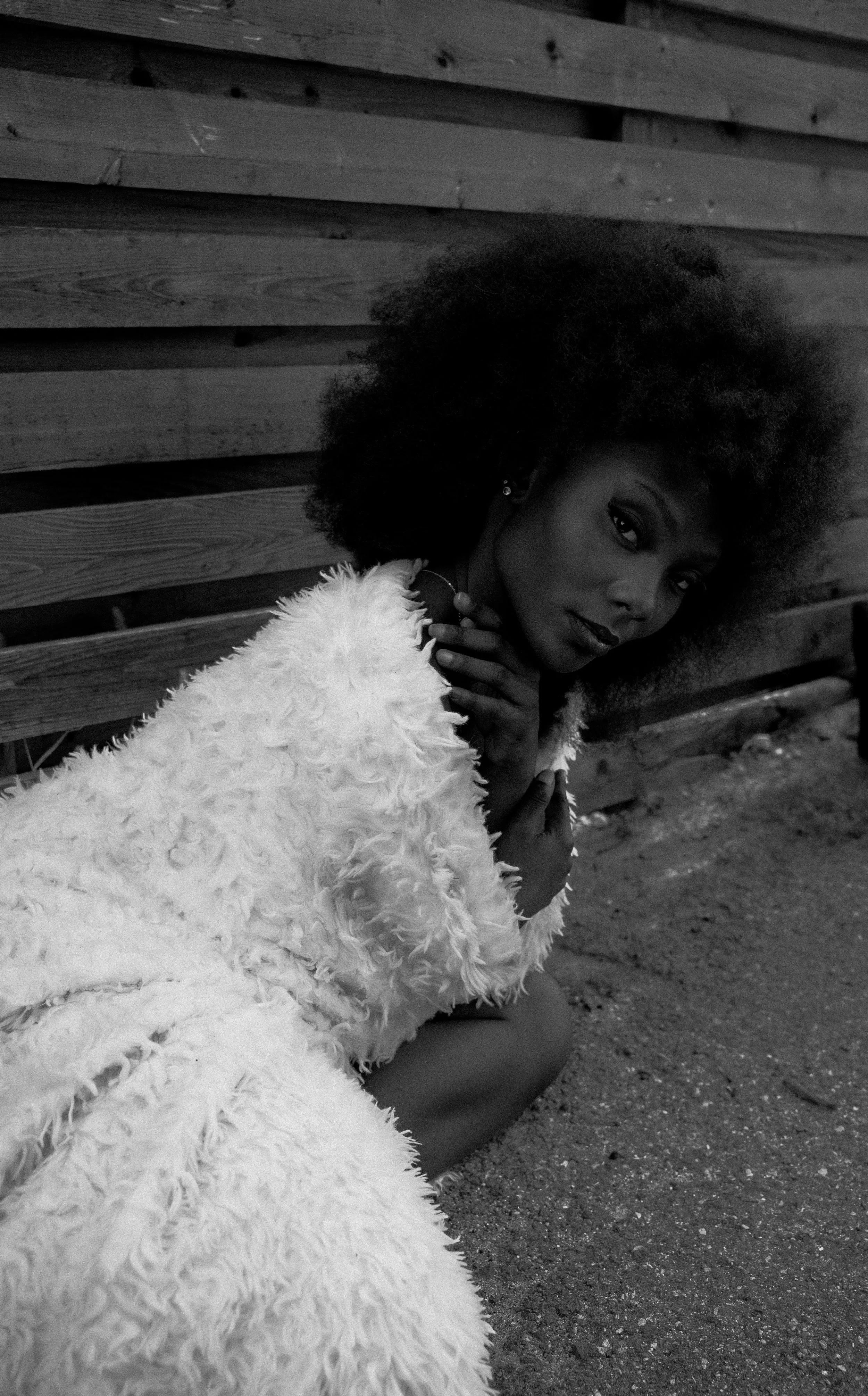 Black and white photo of a woman with voluminous afro hair, sitting on the ground against a wooden fence, wearing a fluffy white coat, looking at the camera with her hands near her face.