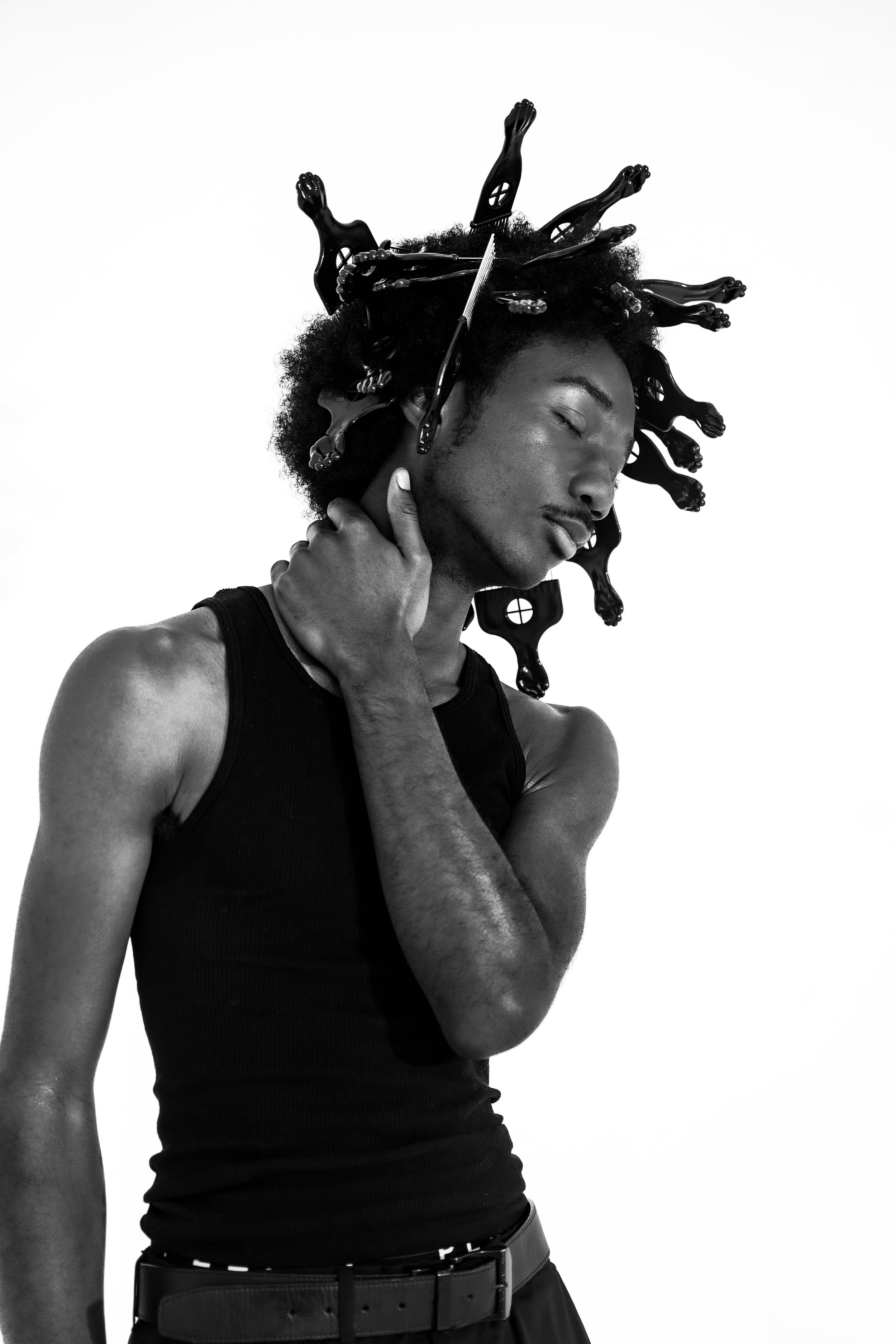 Black and white photo of a young man with medium skin tone, wearing a sleeveless shirt, with hair styled into multiple small braids adorned with black plastic clips. His eyes are closed, and he is touching his neck with one hand.