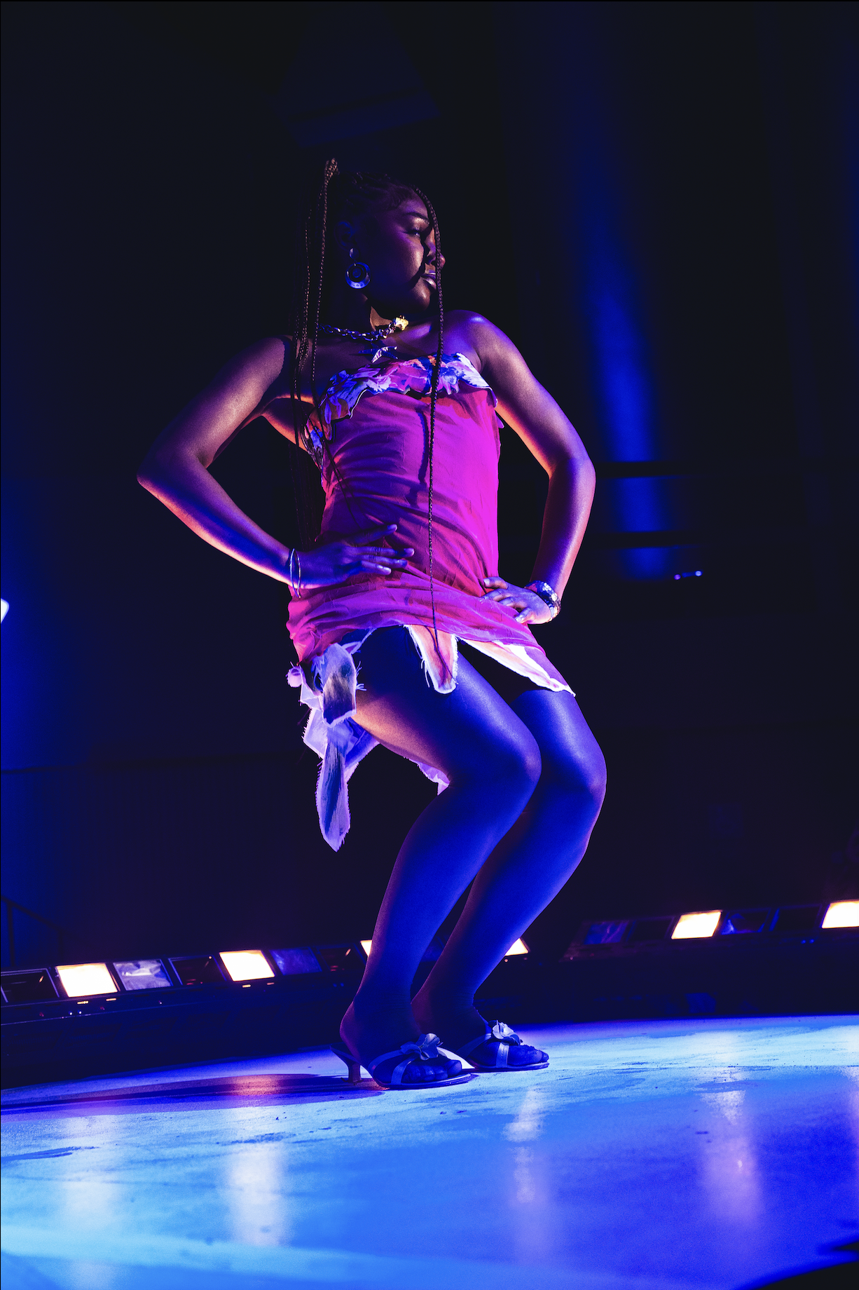 A woman on stage wearing a pink dress with a feathered hem, black stockings, and heels, performing or modeling under stage lights.