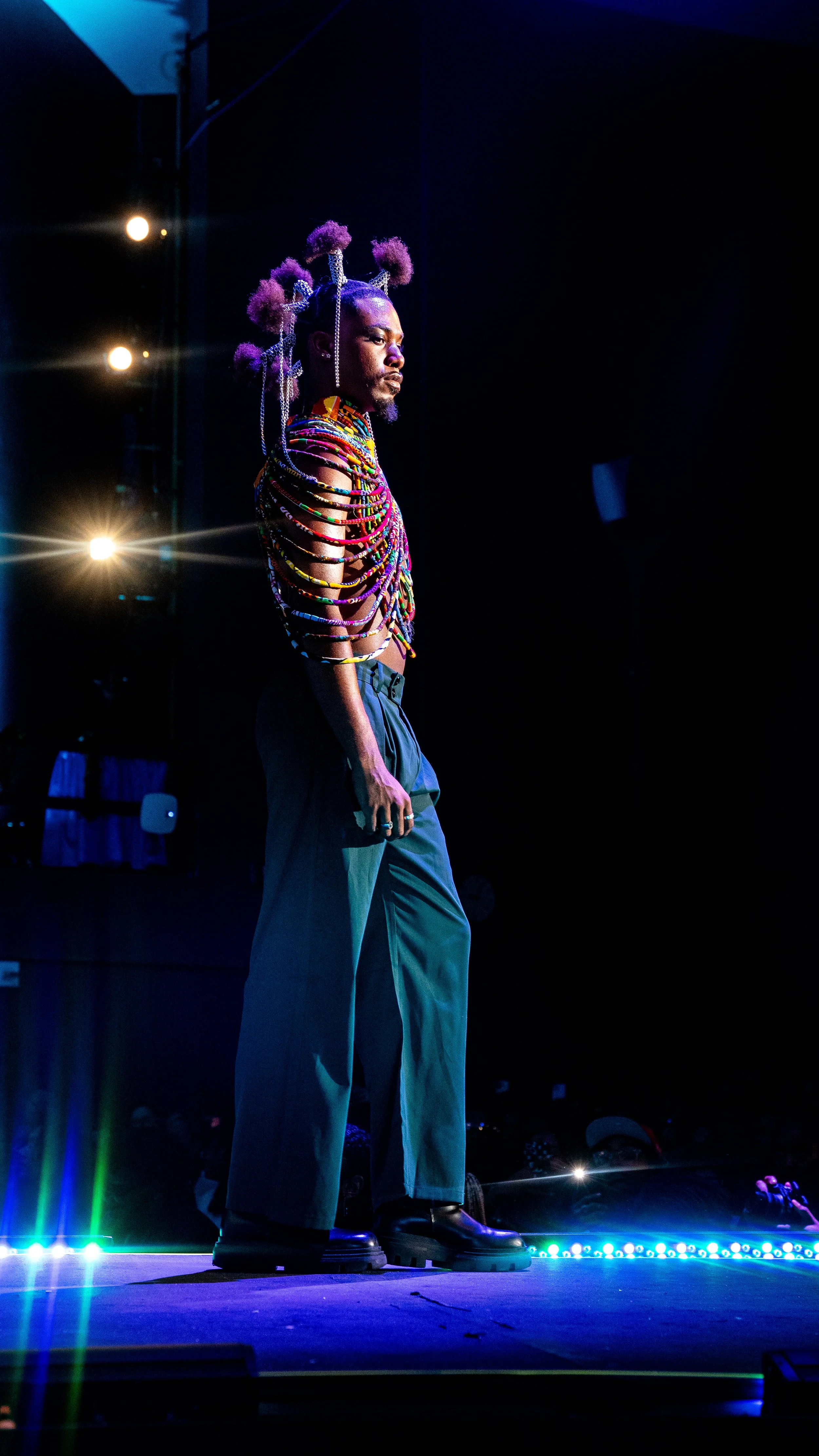A person standing on a stage, dressed in a colorful, beaded outfit with multiple necklaces and elaborate hair accessories, under stage lighting.