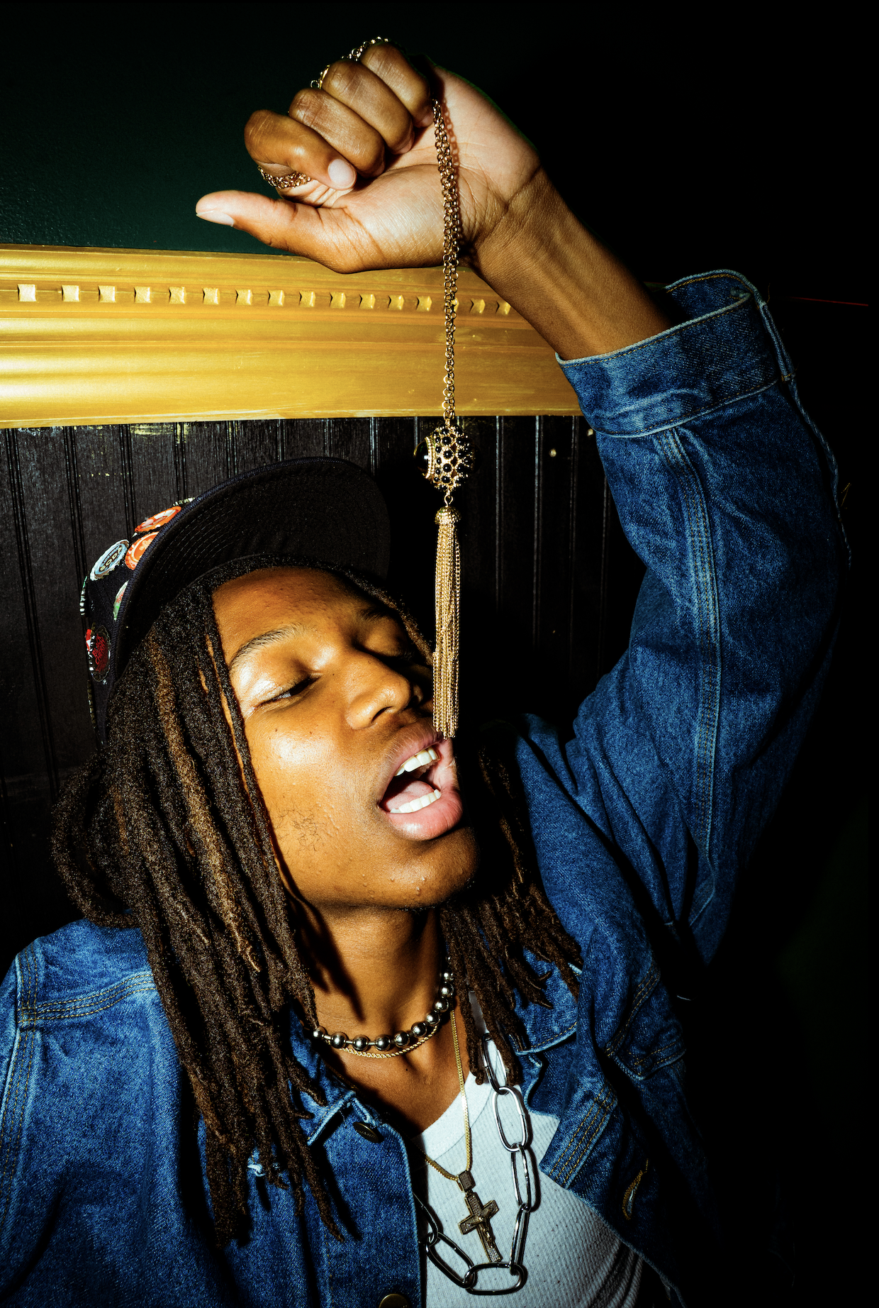 A person with dreadlocks wearing a denim jacket and jewelry, leaning against a dark wall while holding a pendant necklace hanging from their hand, with their eyes closed and mouth slightly open.