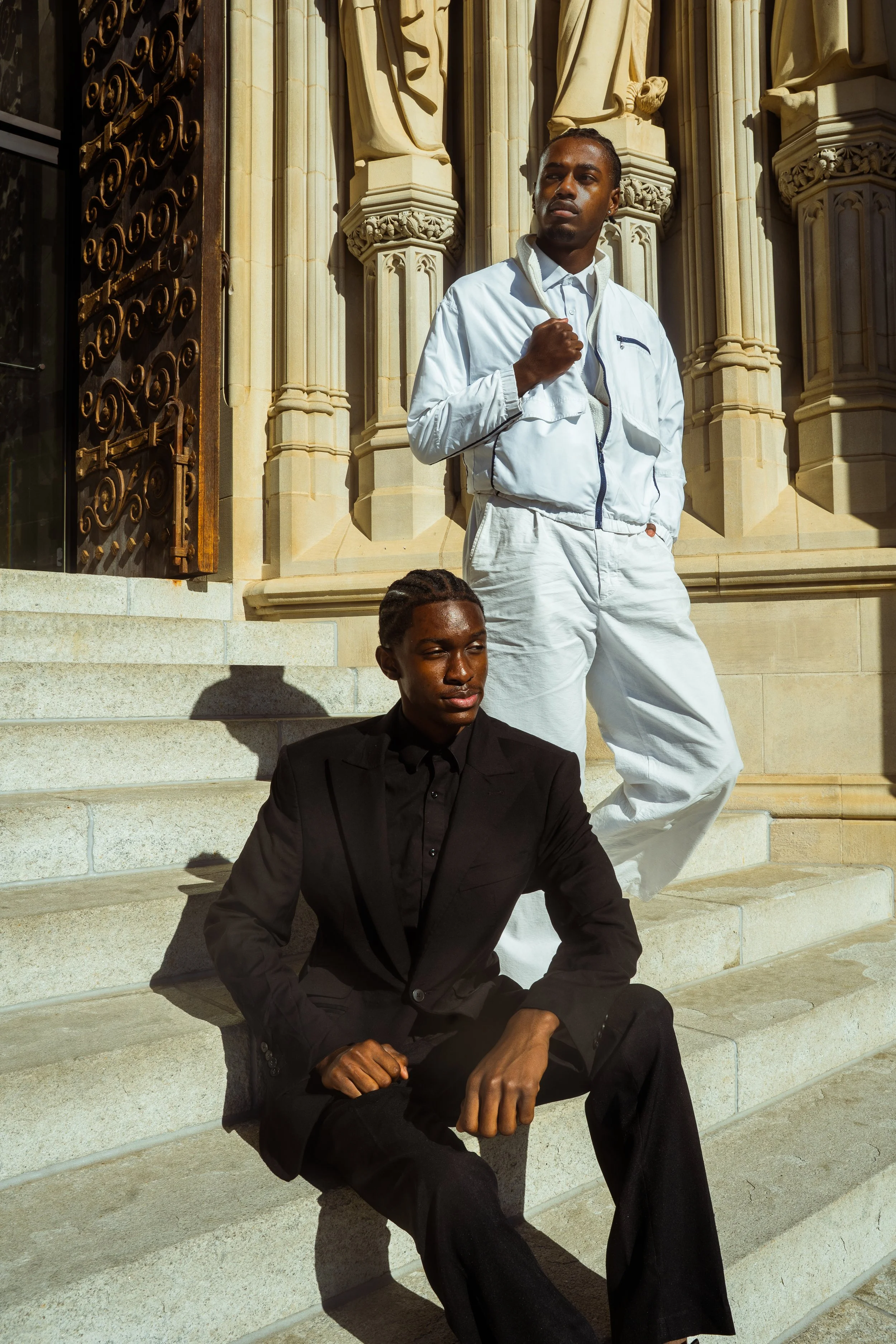 Two stylish young men posing on stone steps in front of a historic building with ornate architectural details. One man is sitting on the steps wearing a black suit, and the other is standing wearing a white tracksuit, both with confident expressions.