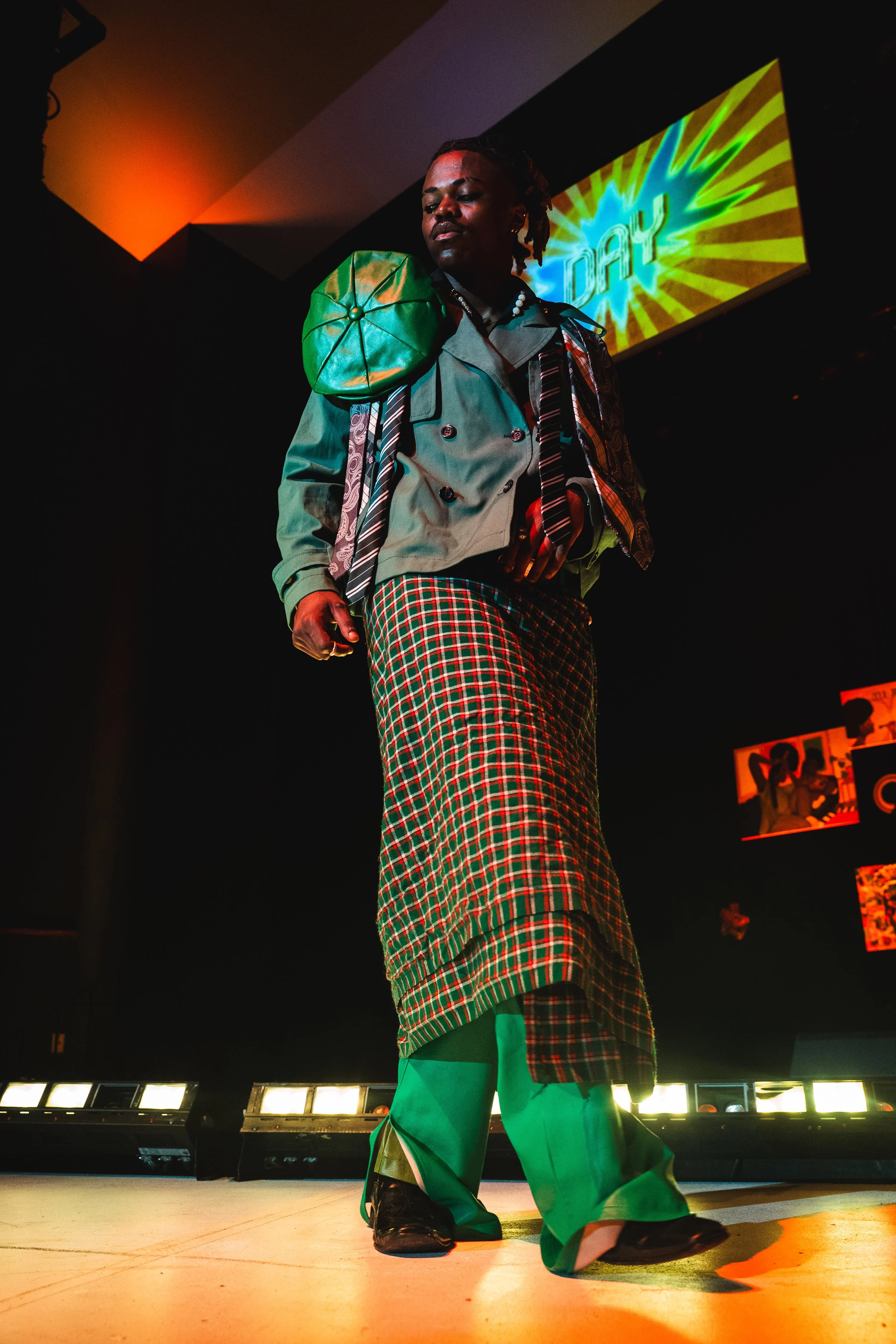 A person walking on a stage wearing a gray trench coat, green pants, and plaid skirt, with a green padded shoulder bag, under colorful lighting with a neon sign reading 'DAY' in the background.
