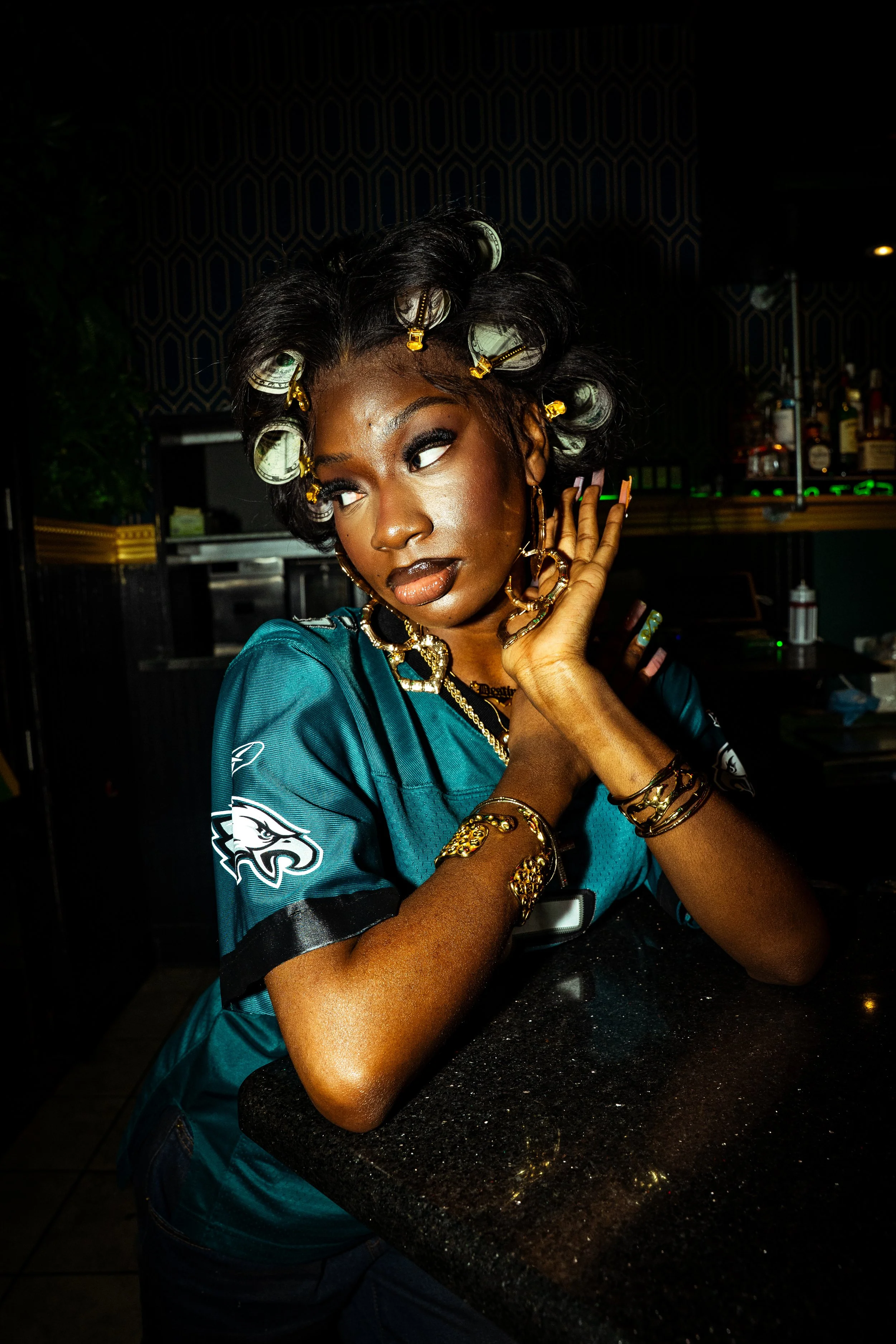 A woman with hair rollers and curlers, wearing a teal Philadelphia Eagles football jersey, and multiple gold jewelry pieces, posing indoors at a bar or restaurant.