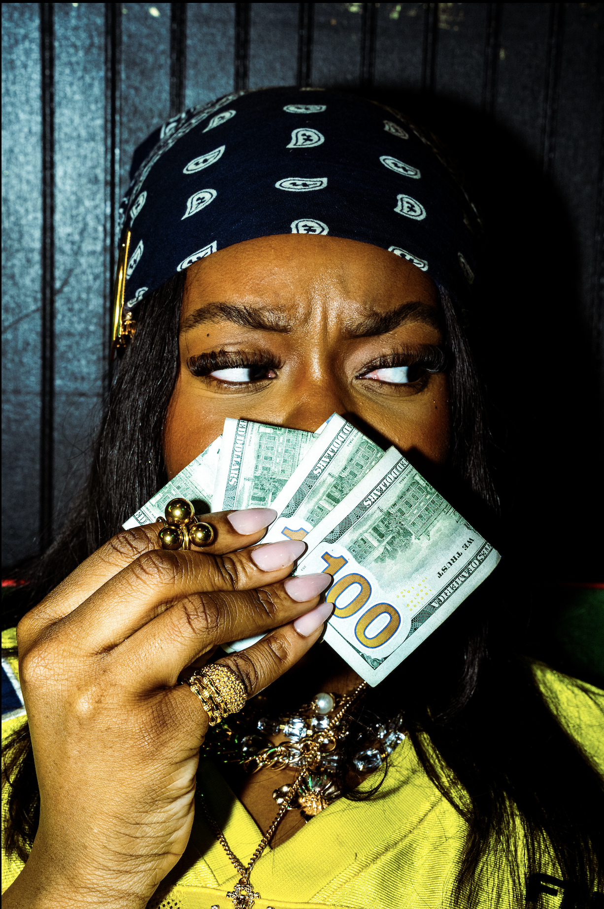 Person with dark hair wearing a blue bandana holds fan of hundred-dollar bills in front of face, showing eyes and jewelry, against dark background.