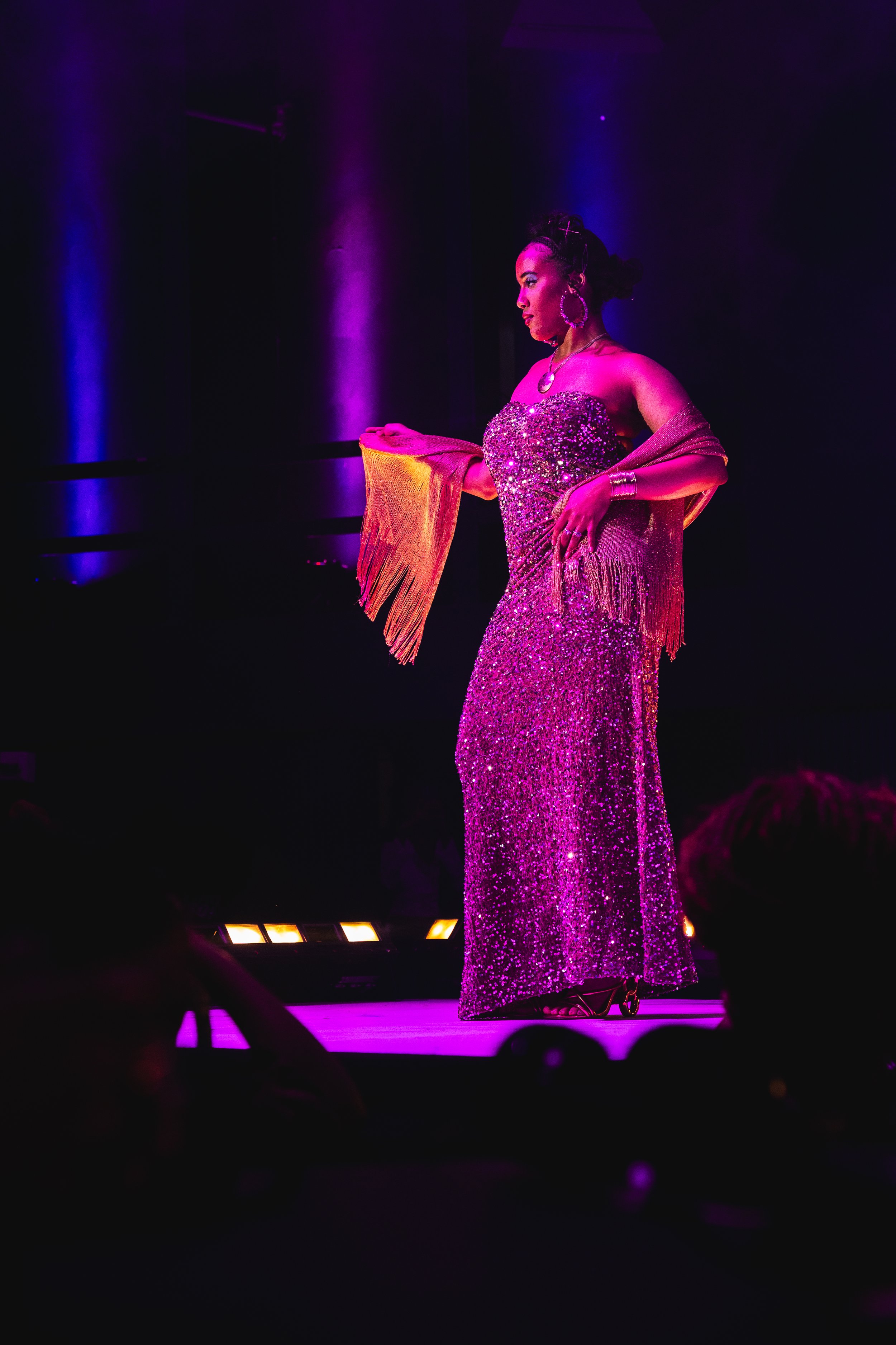 A woman on stage wearing a sparkling, sequin purple gown and matching jewelry, holding a gold fringed shawl, with purple lighting highlighting her silhouette.