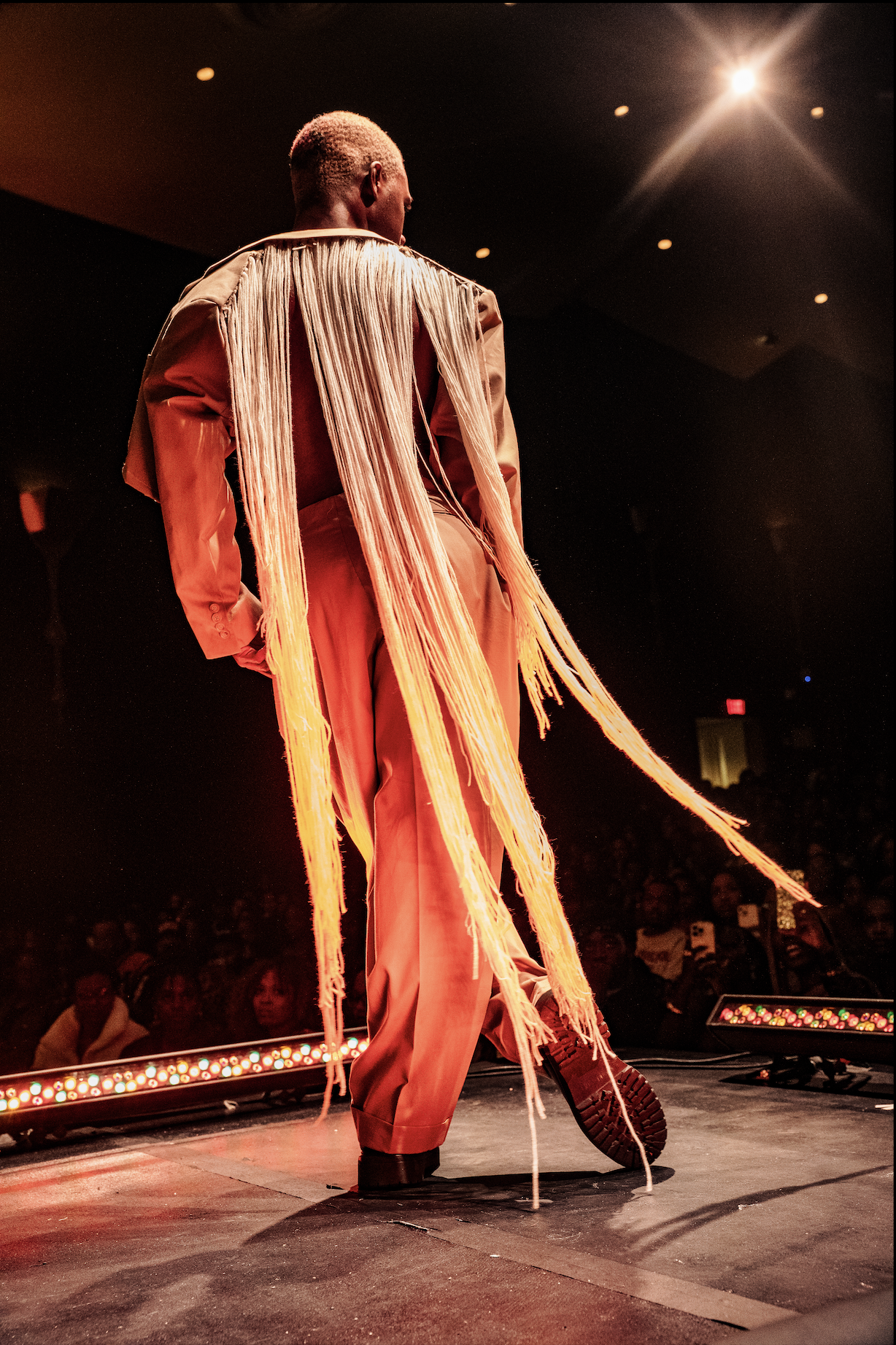 Person wearing a beige pants and jacket with long tassels standing on a stage under bright lights, facing away from the camera, with an audience in the background.