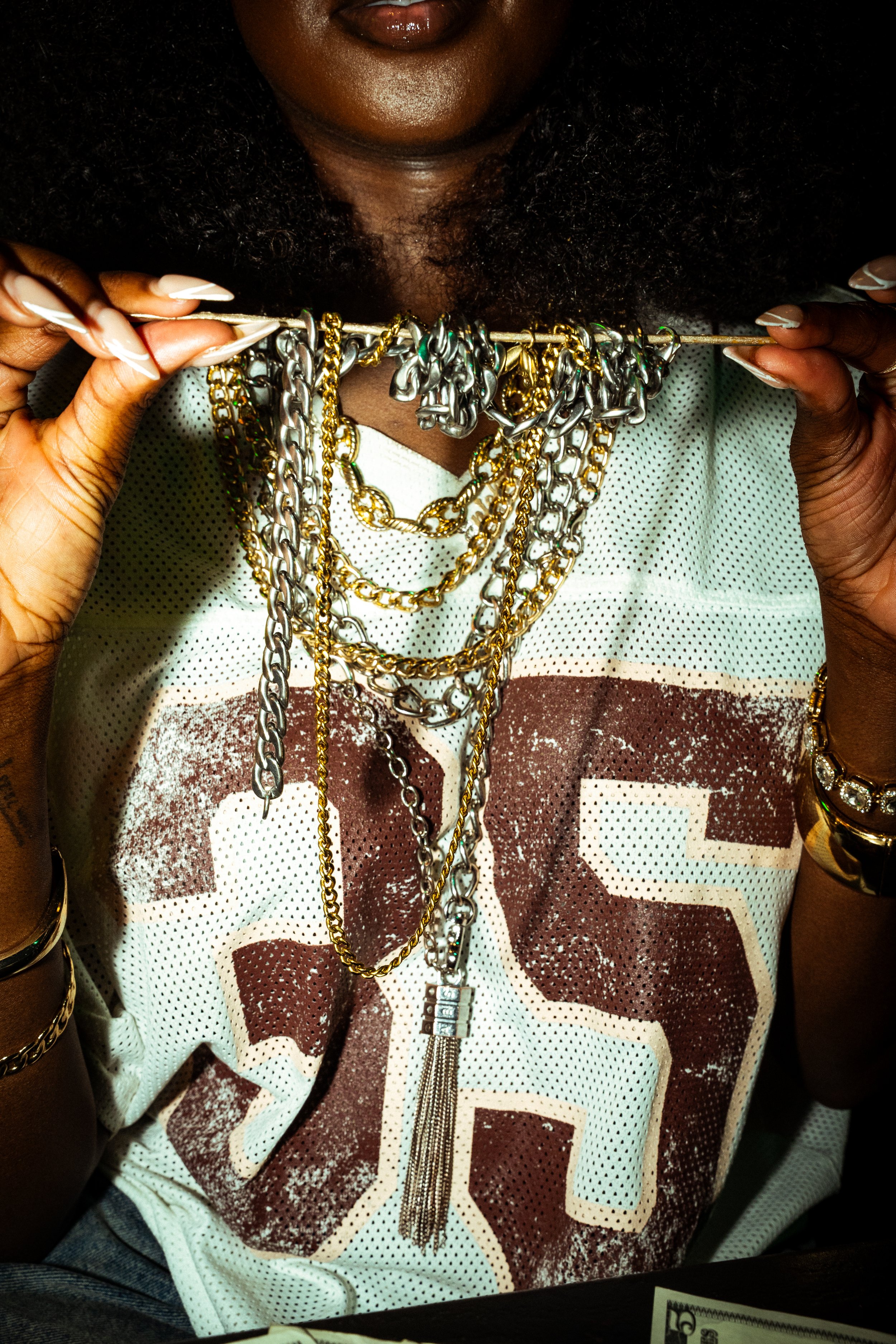Close-up of woman holding multiple gold and silver necklaces on a bar, wearing a sports jersey with a large number 45, with additional jewelry on her arms.