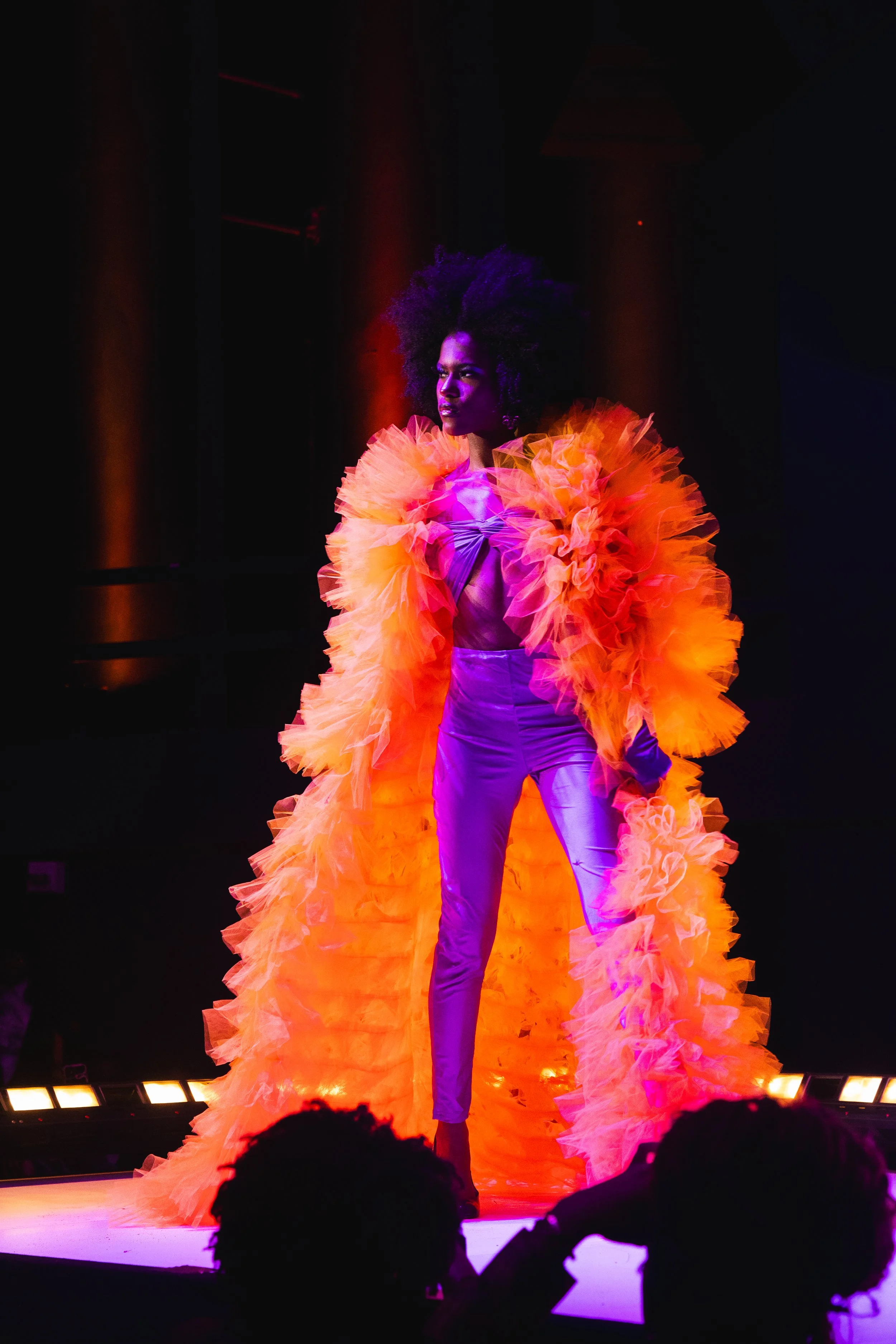 Model on stage wearing a bright orange ruffled coat over purple pants and top, under colorful stage lighting.