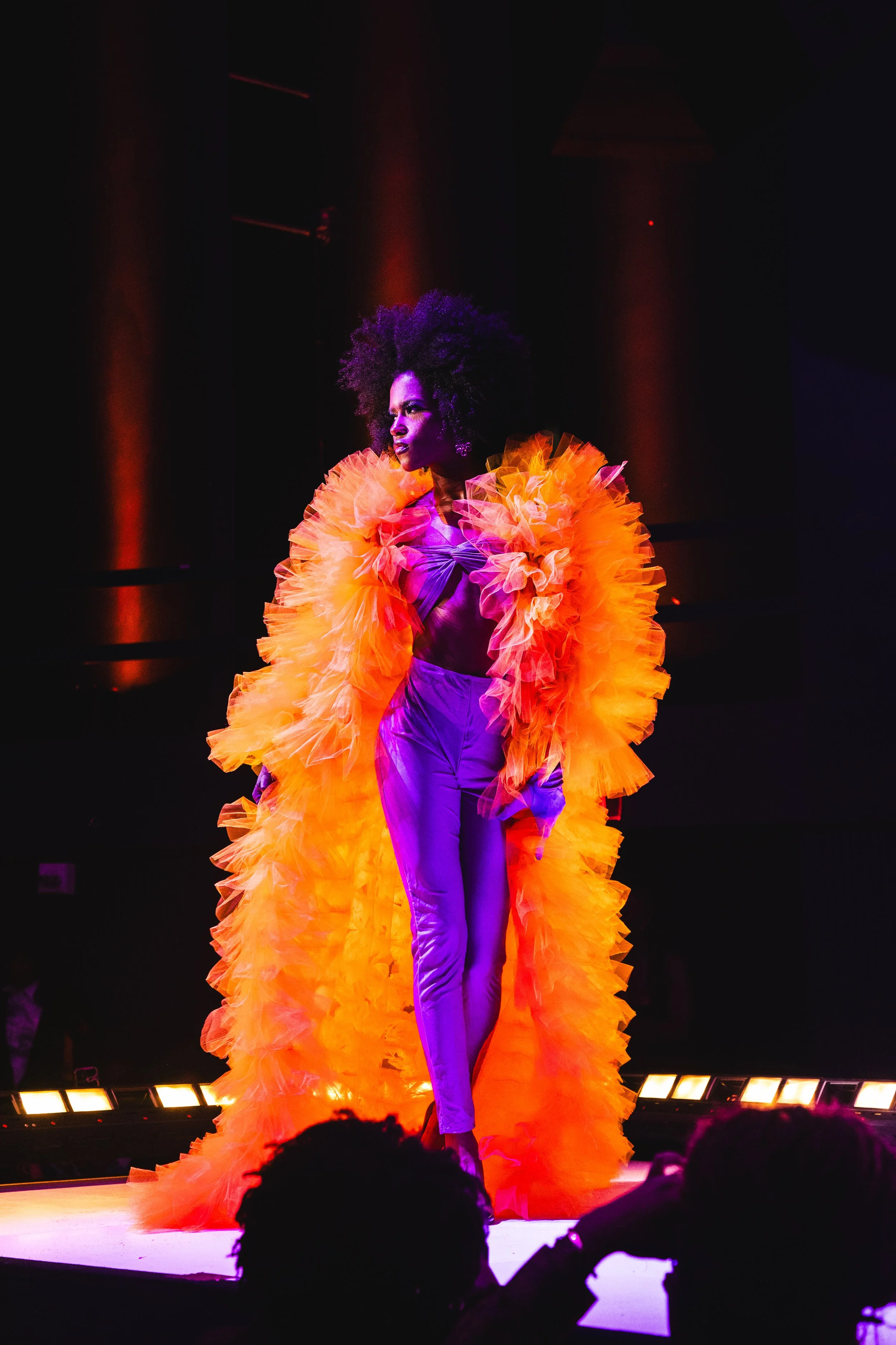 A woman with curly black hair on a stage, wearing an elaborate orange ruffled gown over purple pants, under dramatic lighting.