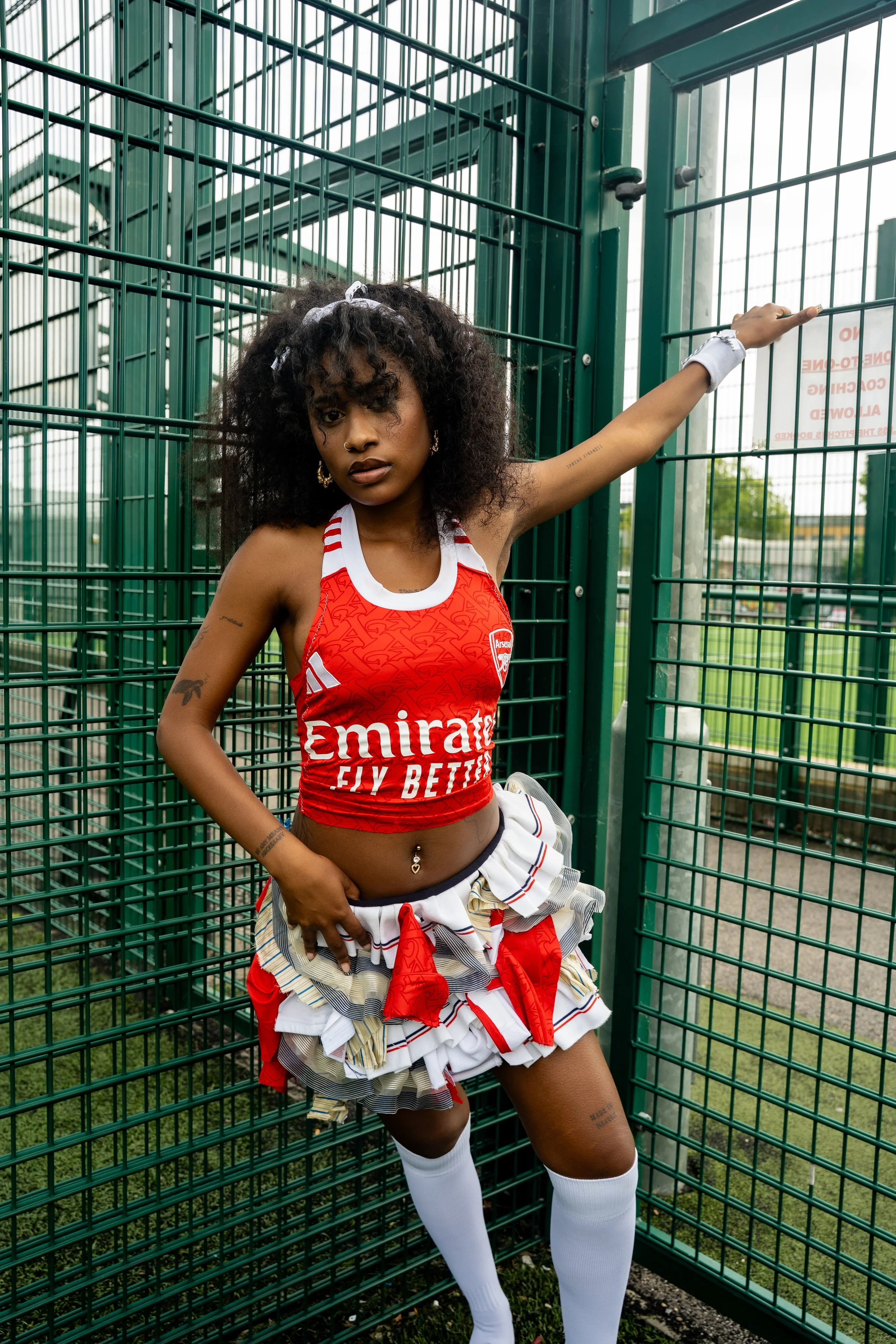 A young woman wearing a red Arsenal football jersey and a layered skirt made of various striped and patterned fabrics, standing inside a green metal cage fence outdoors.