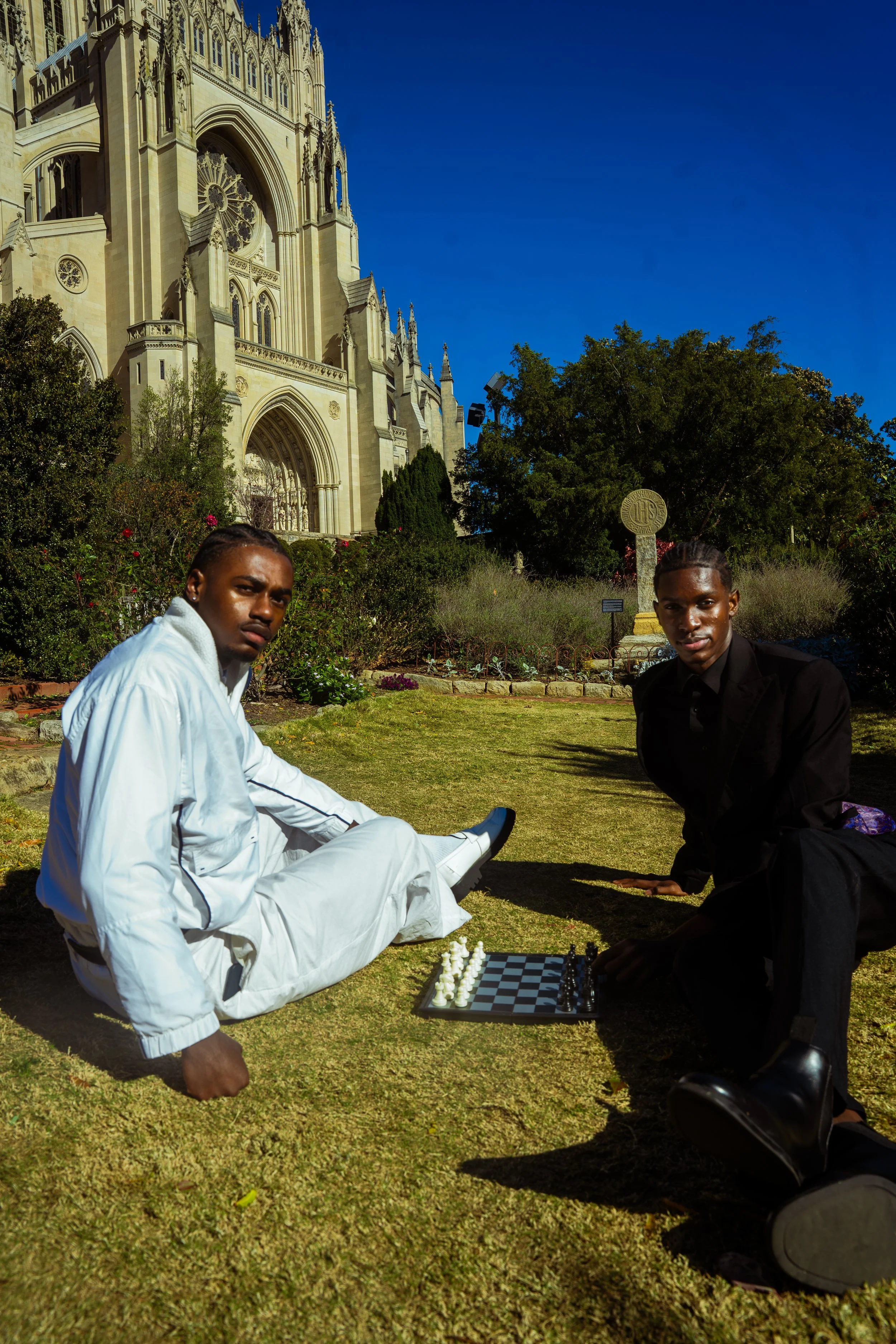 Two young men playing chess on the grass outside a large, gothic-style church surrounded by trees and shrubs, under a bright blue sky.