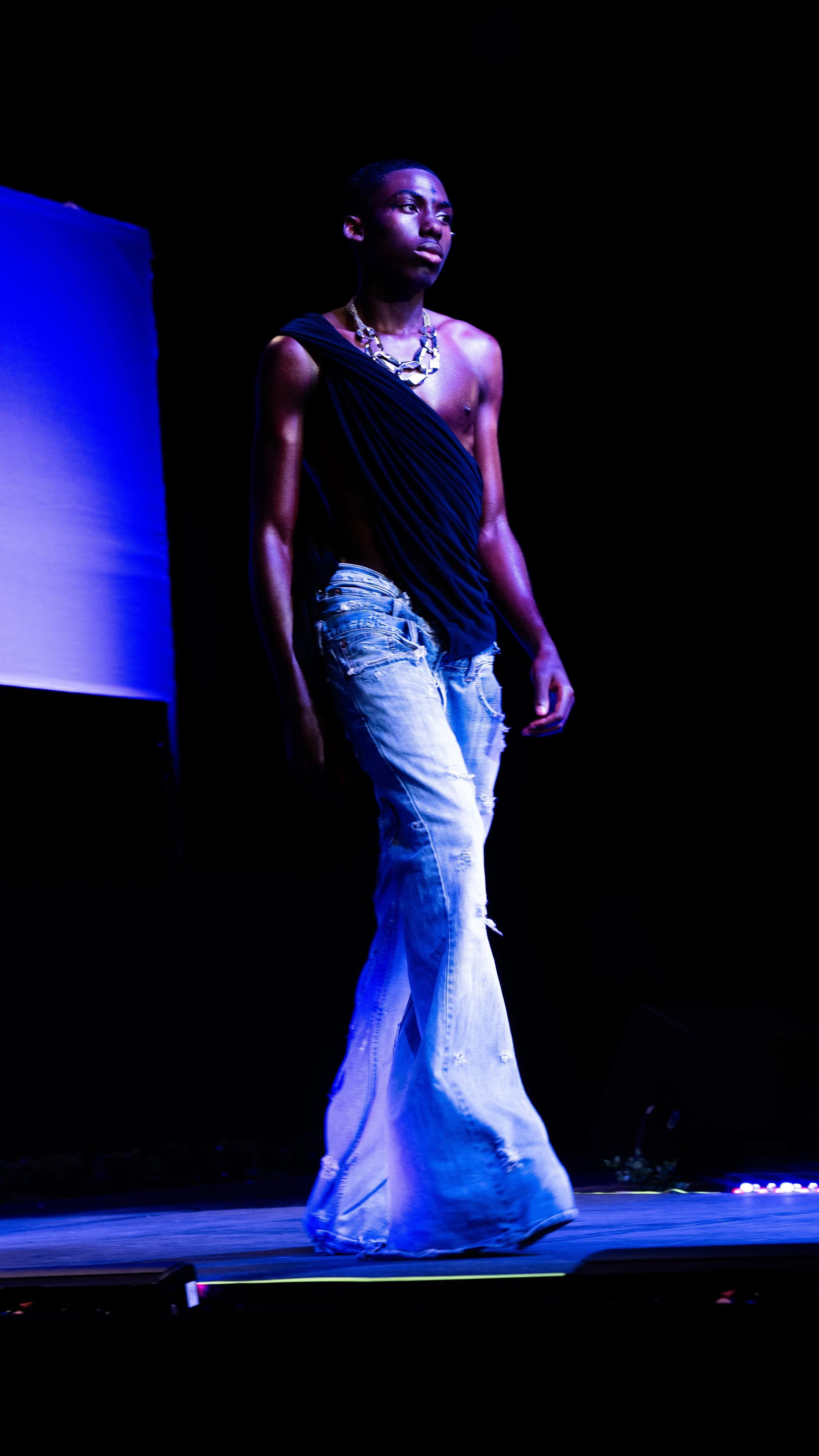 A person with dark skin walking on a runway during a fashion show, wearing a one-shoulder black top and distressed wide-leg jeans, accessorized with a chunky silver necklace, under stage lighting.