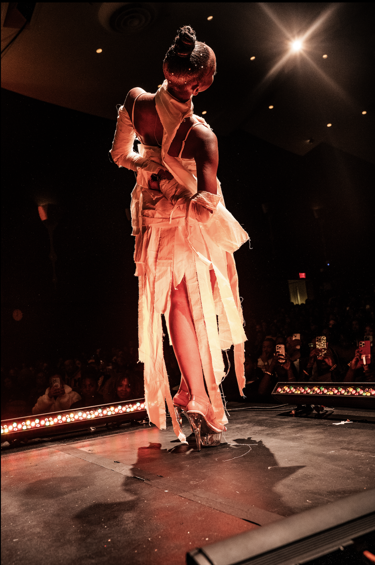 A performer on stage in a dramatic pose, wearing a beige textured dress and clear high heels, with the audience taking photos in the background.