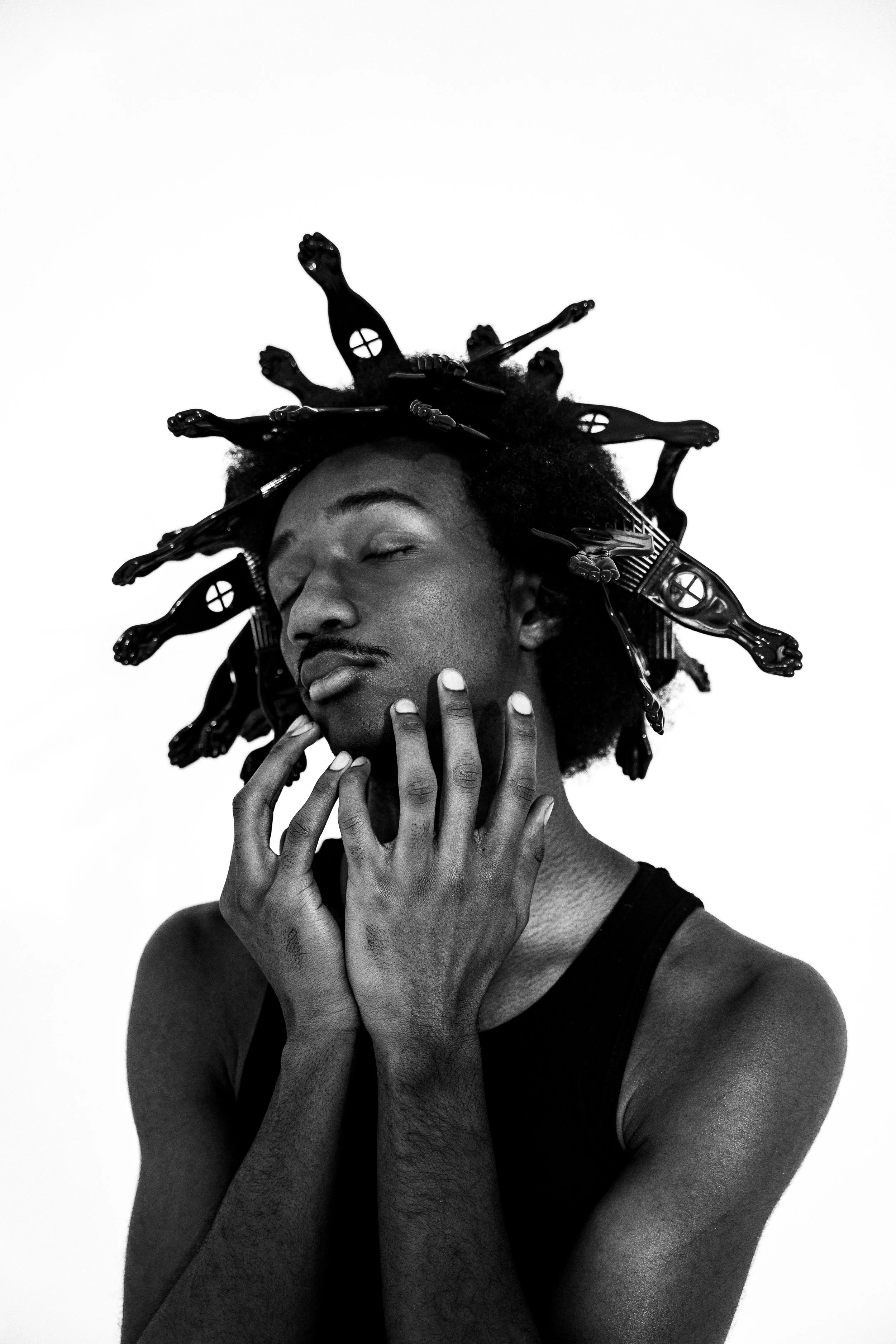 A black and white photo of a man with his eyes closed, touching his face with his hands. His hair is styled with multiple miniature dollhouses attached, creating a stencil-like effect.