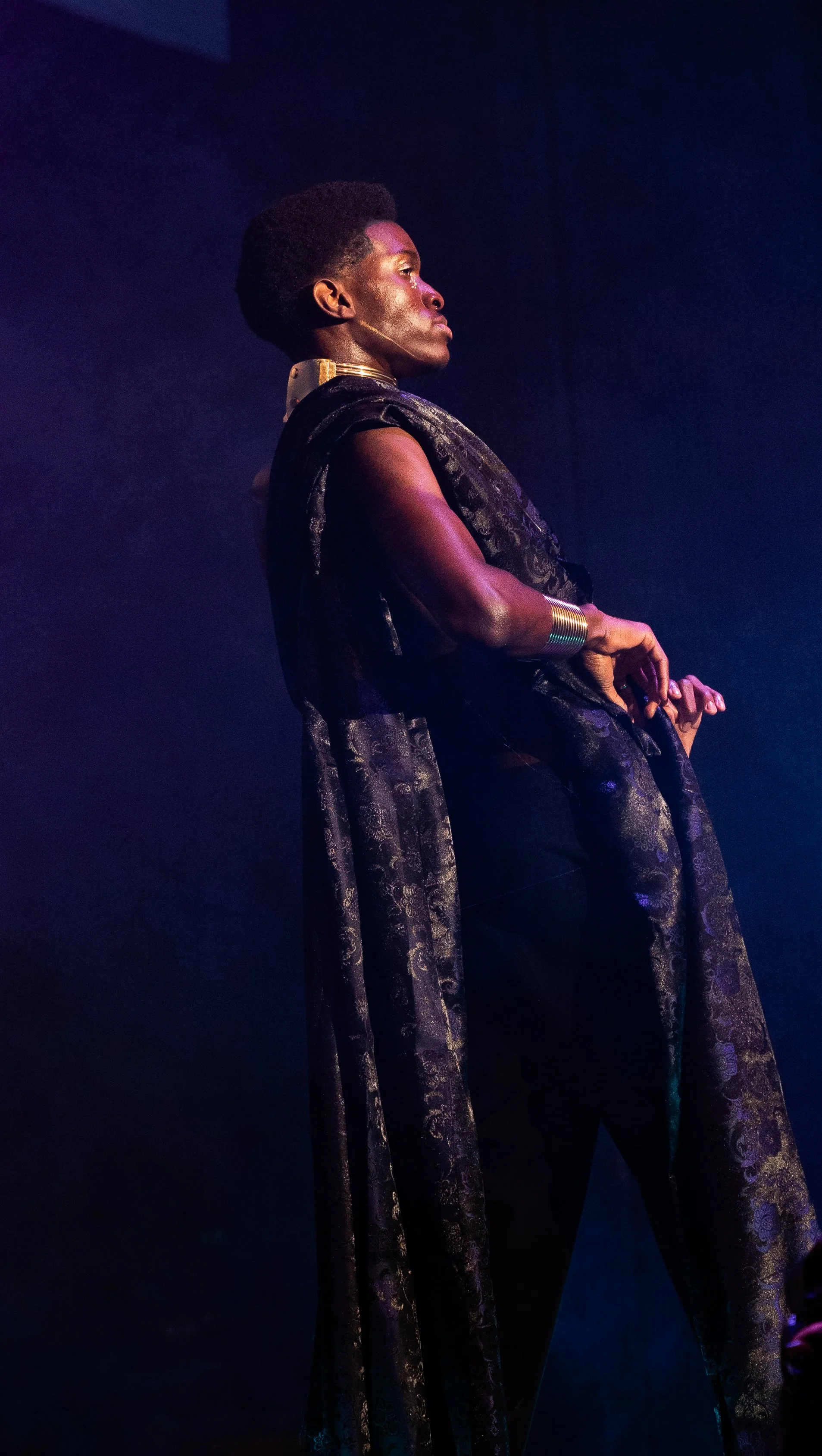 A person in elaborate black and gold costume with dramatic makeup performing on stage with blue lighting.