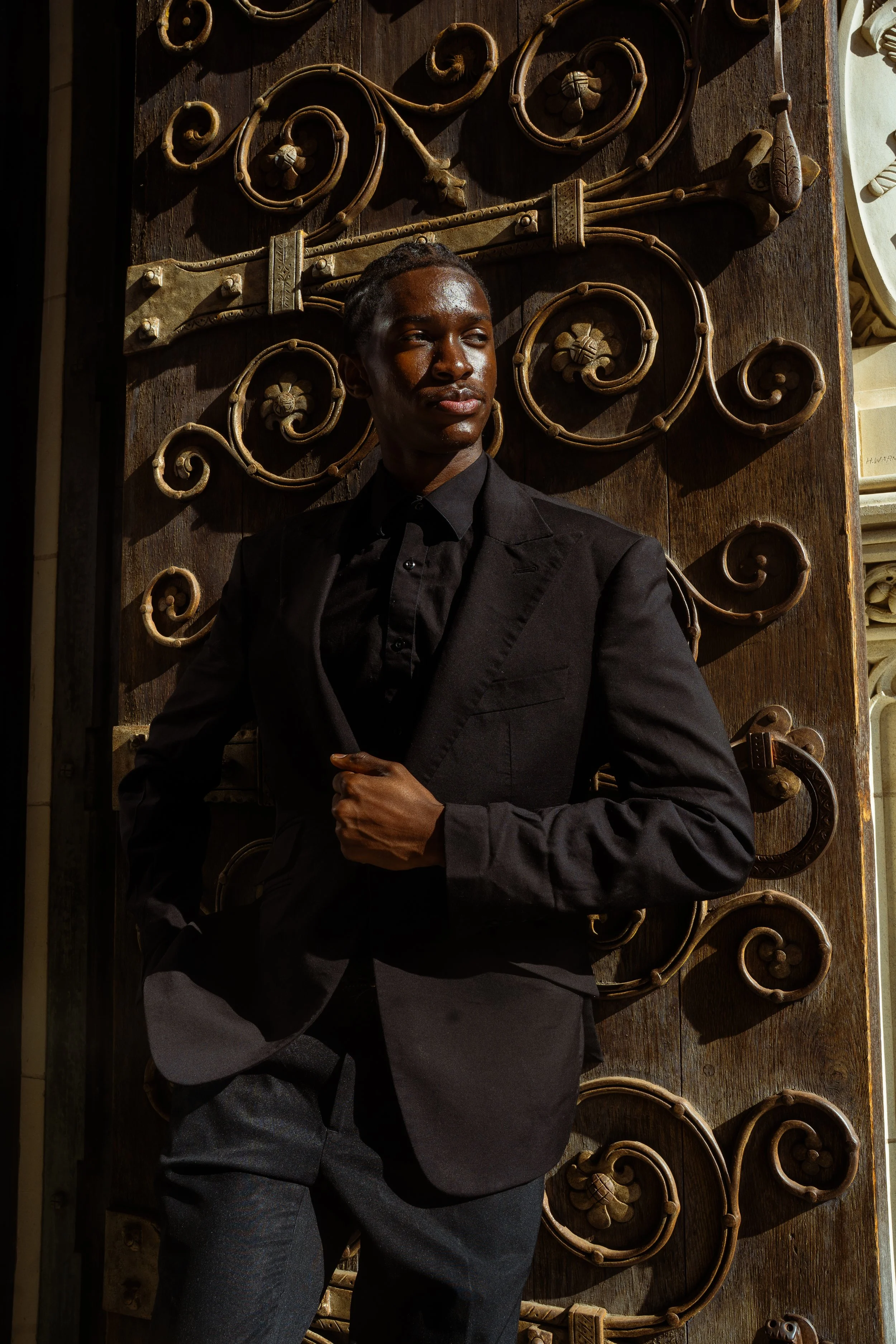 A person with dark skin, wearing a black blazer and shirt, standing in front of a decorative wooden door with intricate metal scrollwork, bathed in warm sunlight.