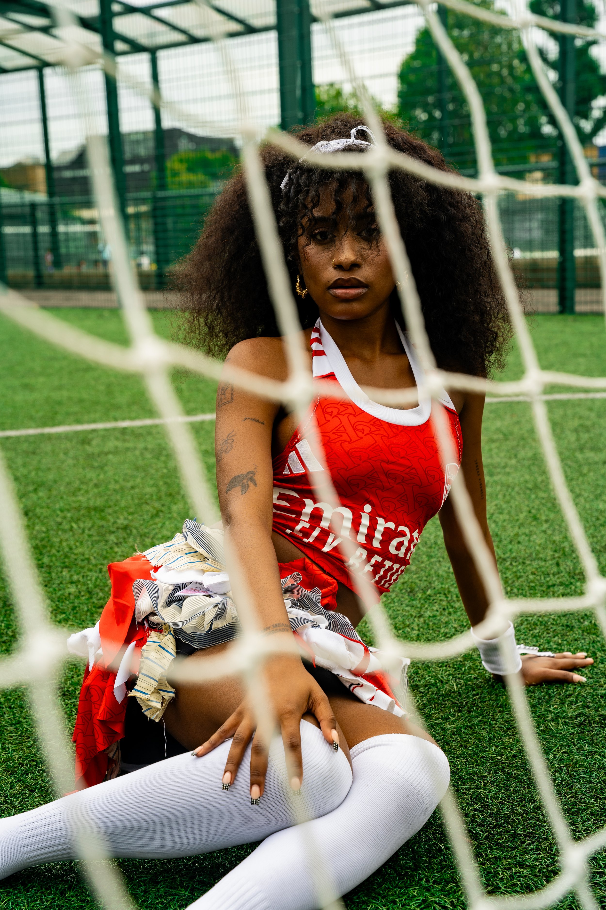 A woman in a red sports outfit with white knee-high socks, sitting on the grass behind a soccer goal net on a field.