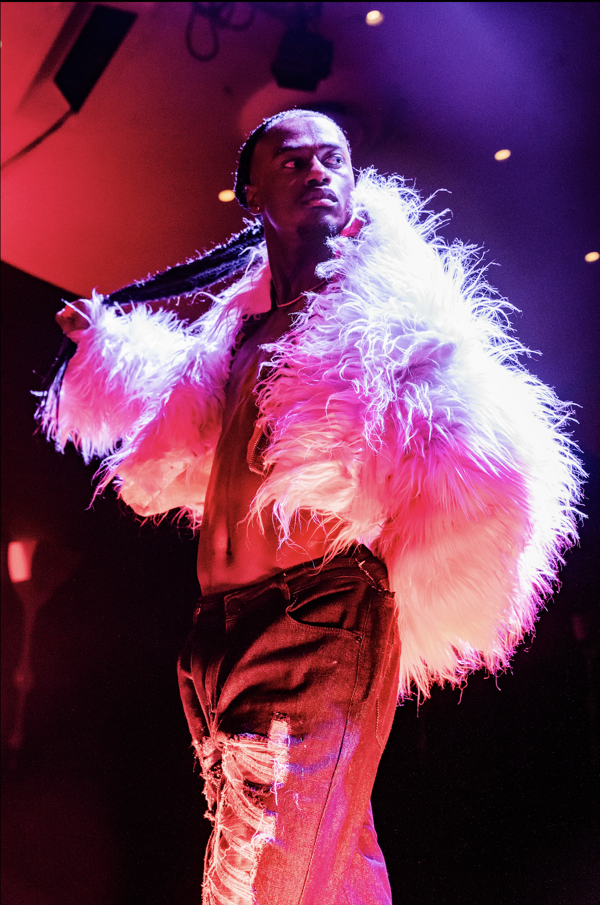 A person wearing a white fur coat and distressed jeans performing on a stage with colorful lighting.