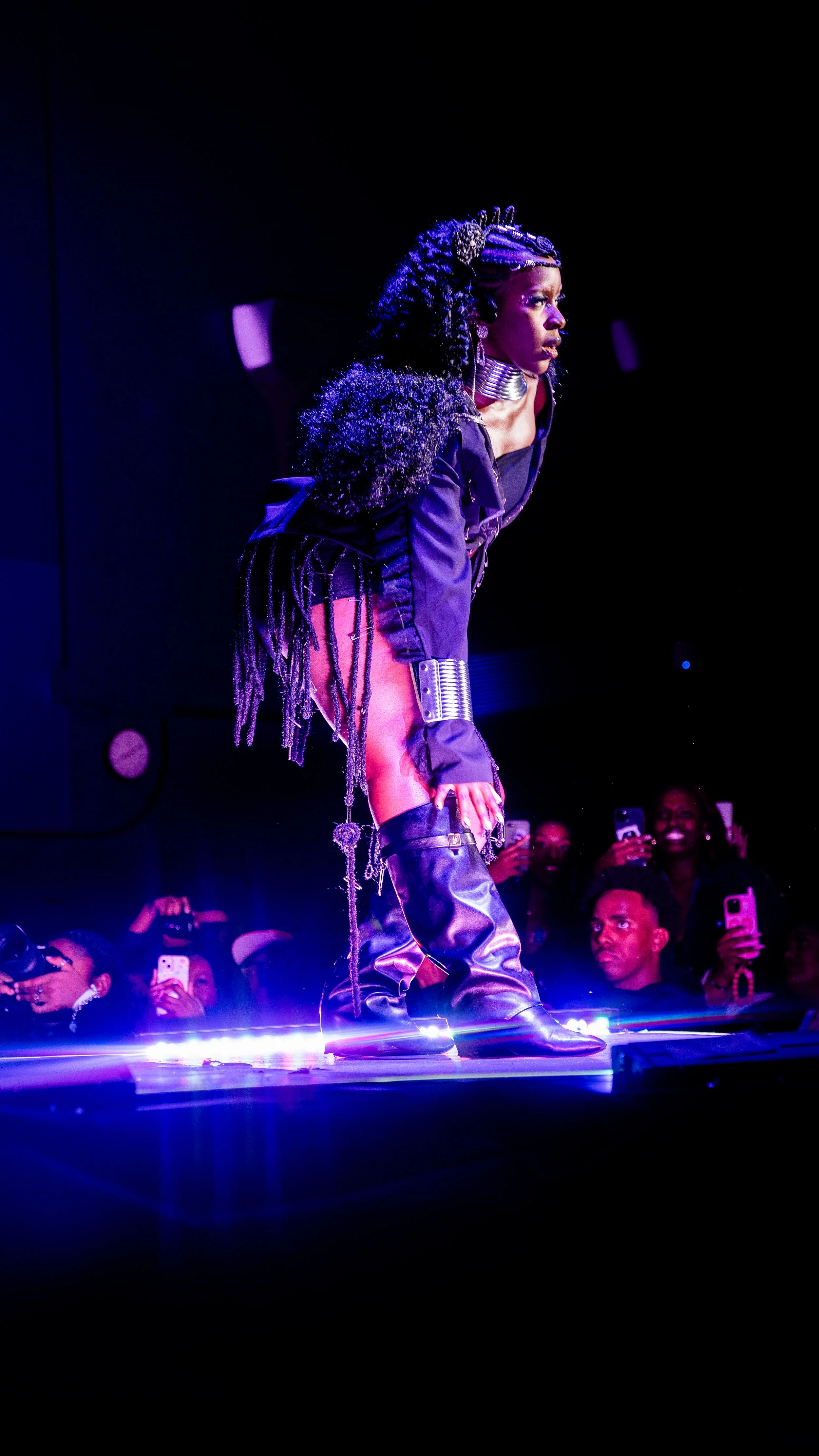 A woman performing on a stage with dramatic lighting, dressed in a dark, edgy outfit with thigh-high boots and silver accessories, surrounded by an audience taking pictures.