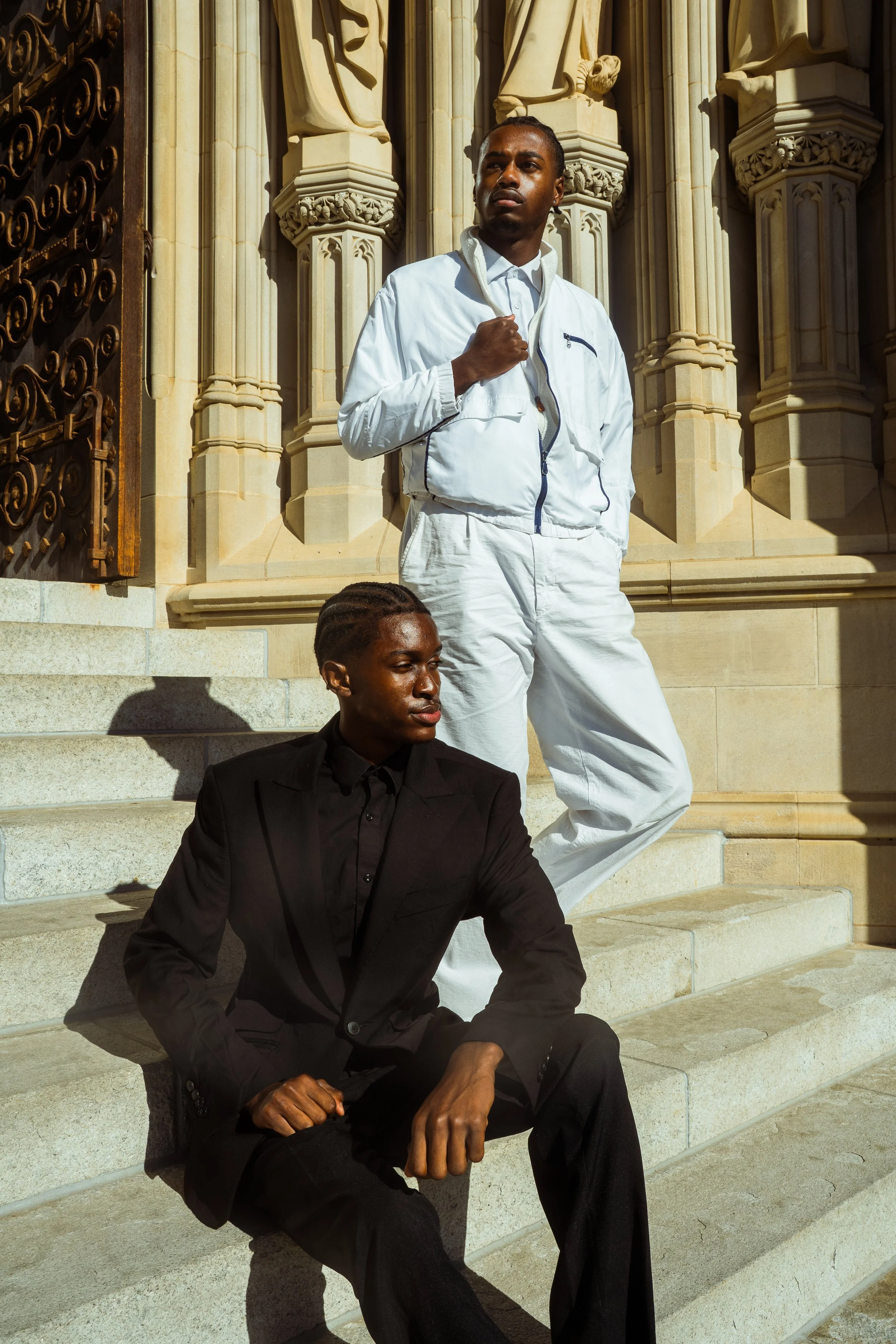 Two young men in stylish outfits pose on stone steps in front of an ornate, Gothic-style building. One is sitting wearing a black suit, and the other is standing in a white tracksuit.