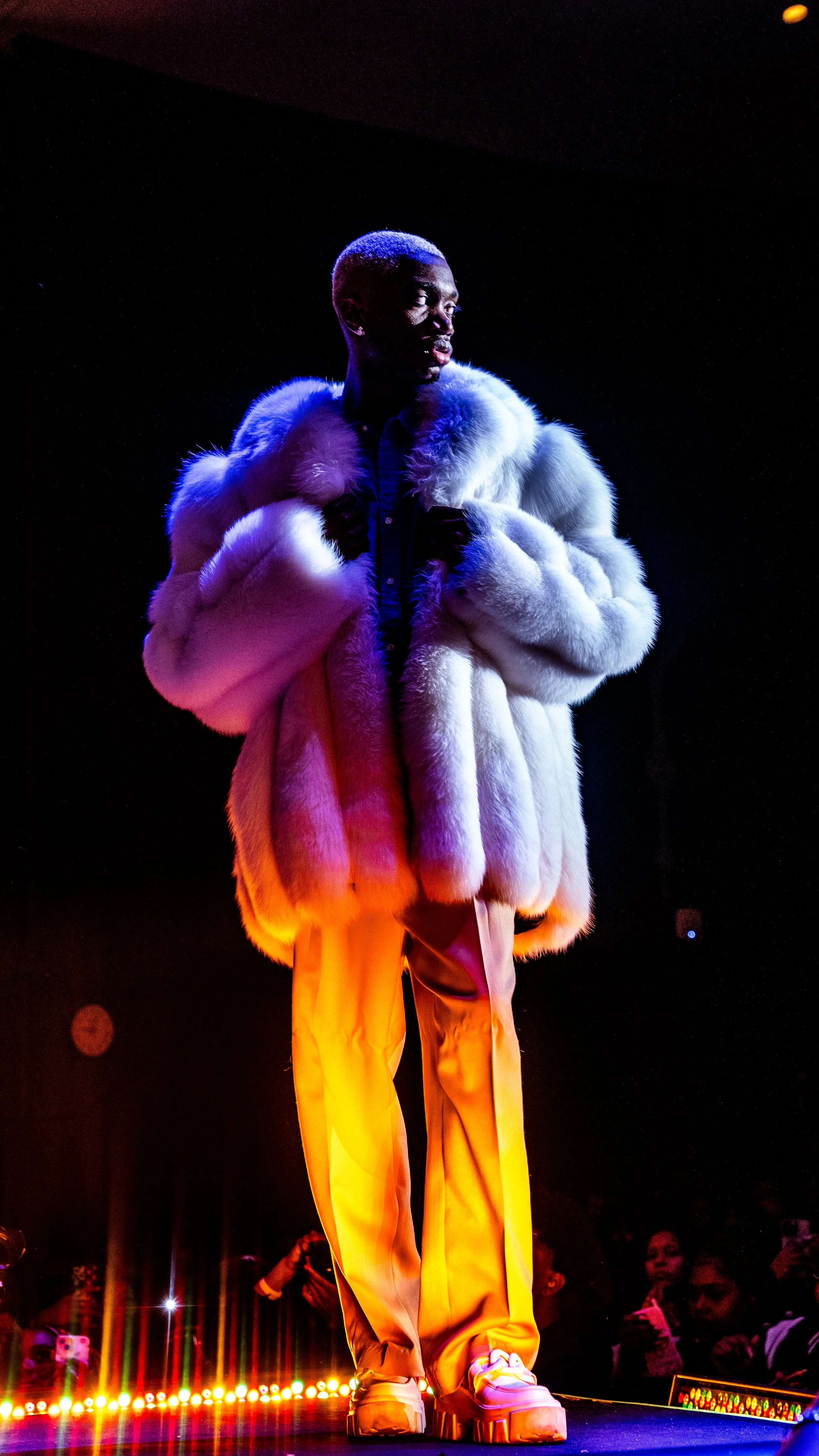 Person posing on stage wearing a white fur coat, yellow pants, and white shoes, with audience members visible in the background under colorful lighting.