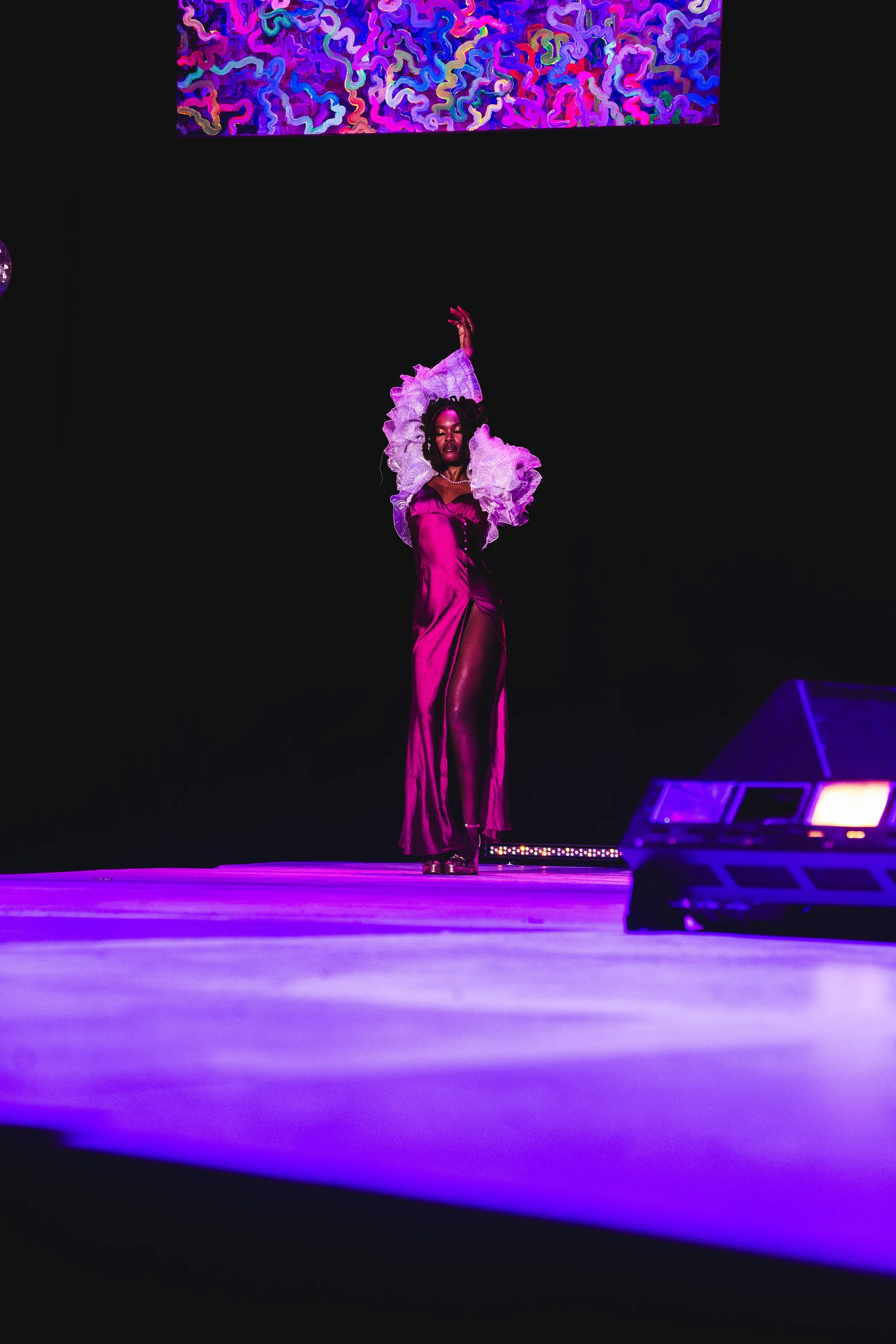 A woman in a shiny pink dress with ruffled sleeves walking on a runway under purple lighting with a dark background, with a colorful abstract artwork above her.