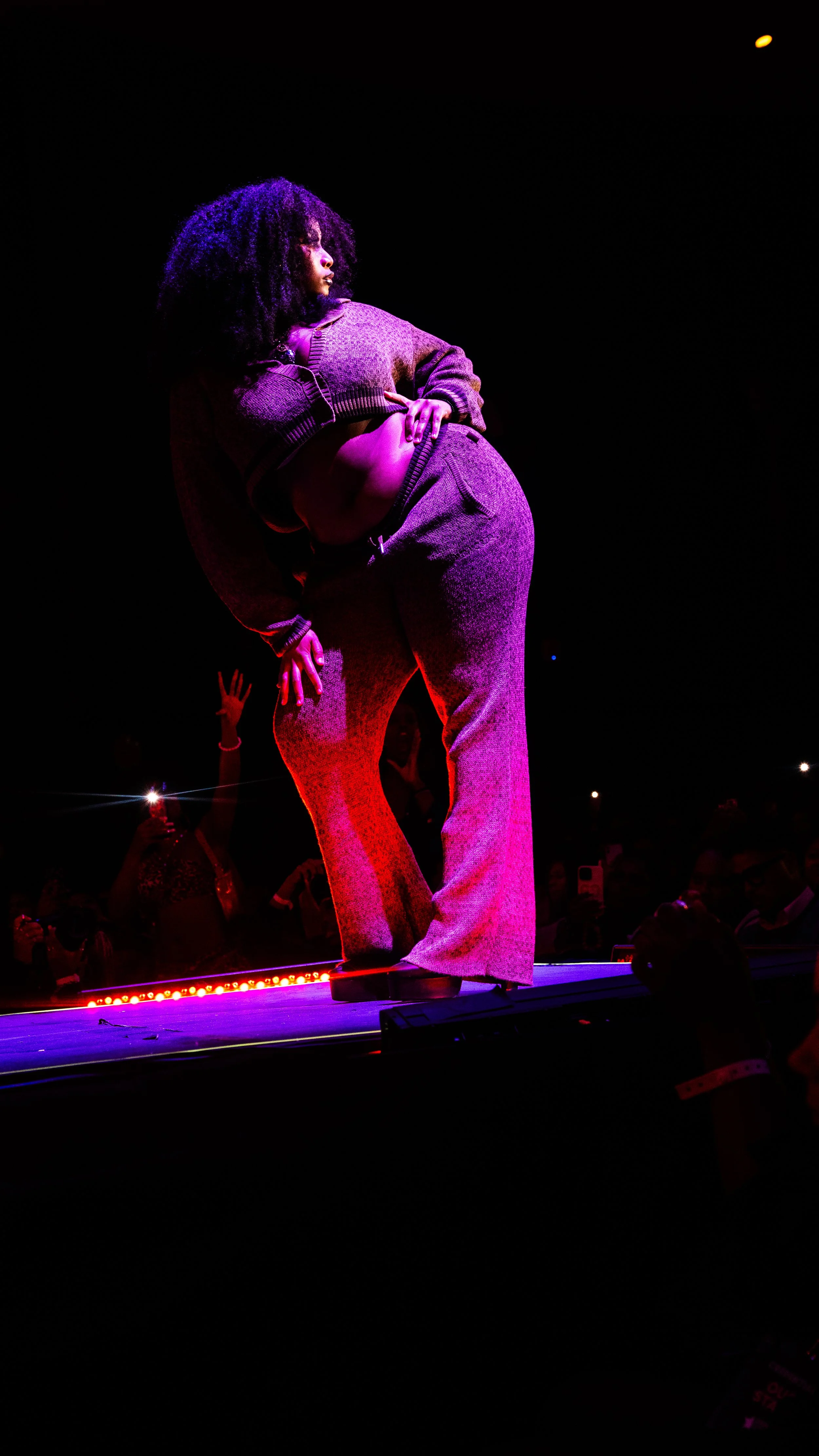 A woman with curly hair standing on a runway stage at night, illuminated by purple and pink stage lights, with an audience in the background.