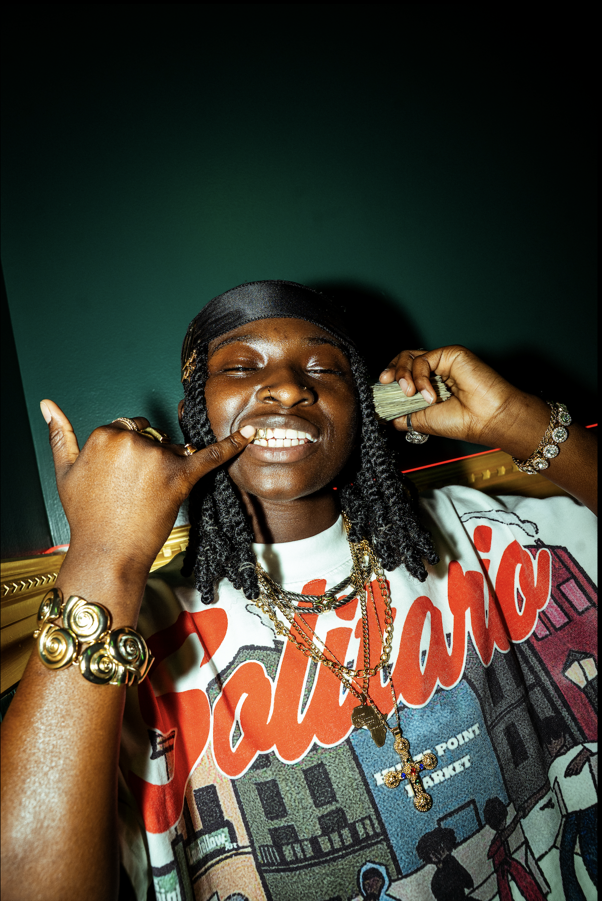 A person with dark curly hair, wearing jewelry including rings, bracelets, and layered gold chains, smiling brightly and making a rock on gesture with one hand near their face. They are wearing a white T-shirt with a colorful graphic and red text.