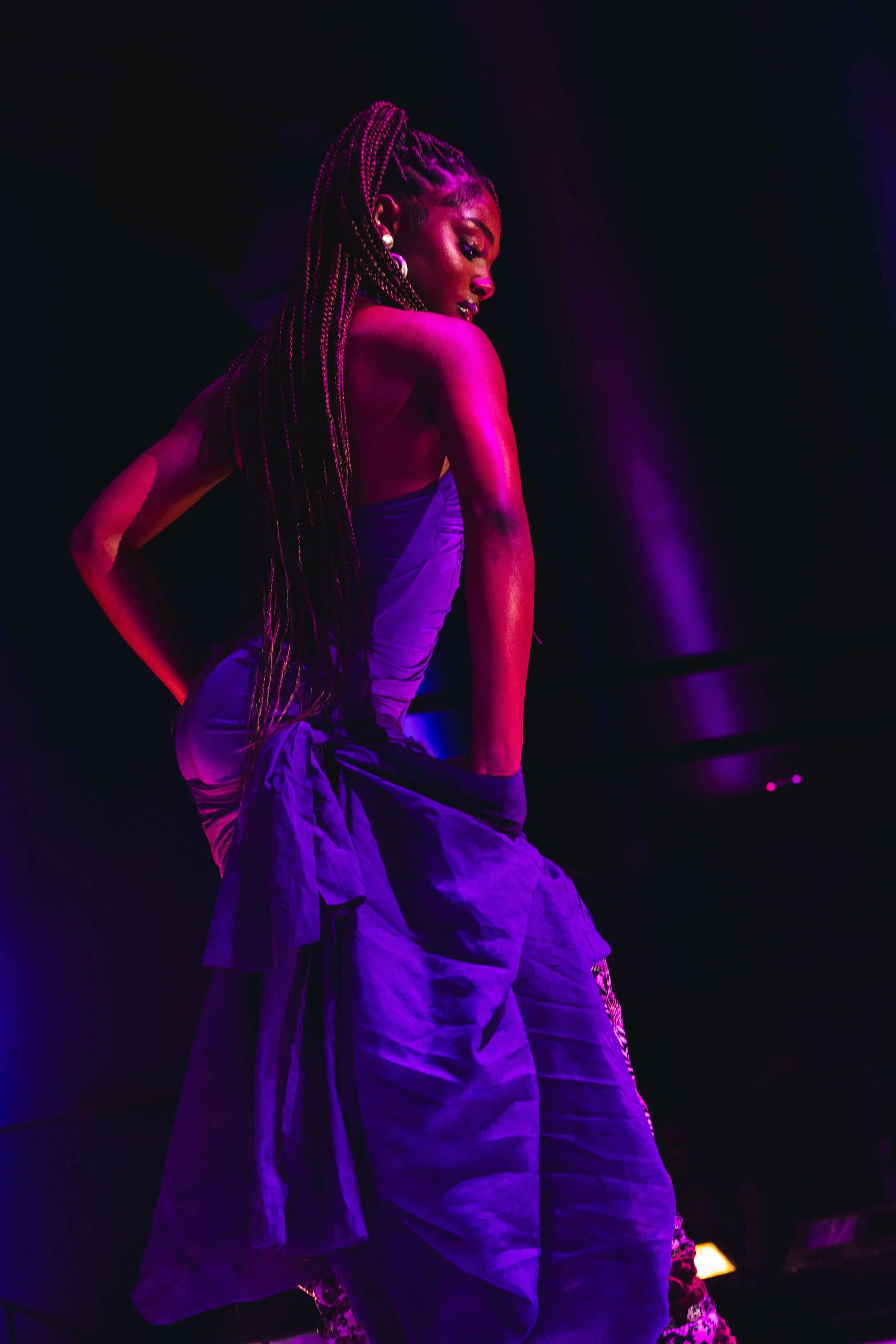 A woman with long braids wearing a purple dress and large earrings, illuminated by pink and purple stage lighting, on a dark stage.